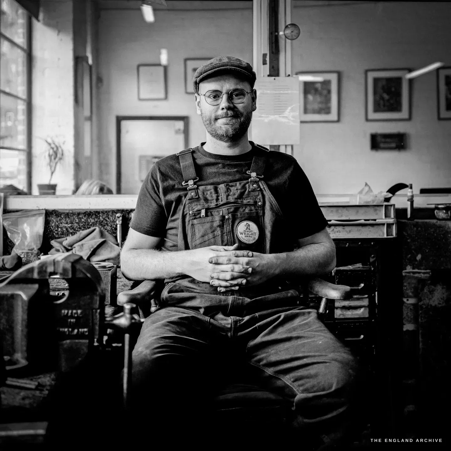 A seated portrait of Sam at the bench, hands clasped, flat cap and glasses, looking to camera, framed prints on the wall behind.