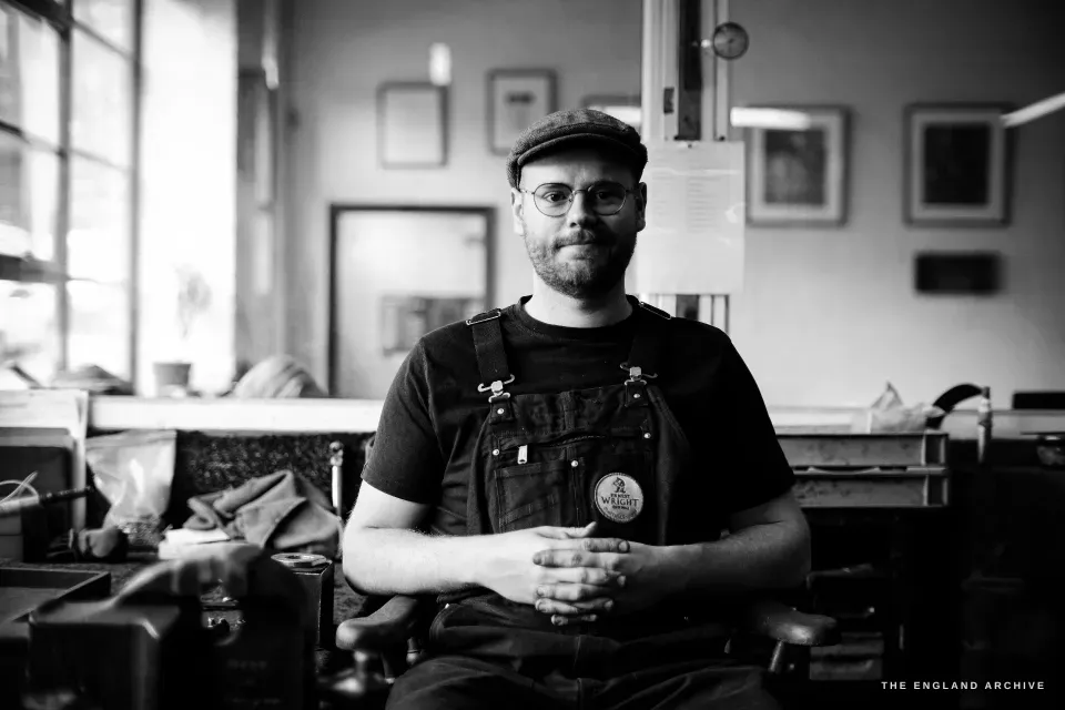 A formal seated portrait of Sam Aston-Clark facing the camera, flat cap, glasses and leather apron, the workshop windows behind him.