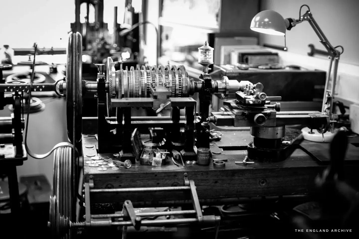 The whole rose engine on its wooden bench - the horizontal stack of patterned cams, the lathe headstock and an anglepoise lamp, watch parts scattered on the bed.