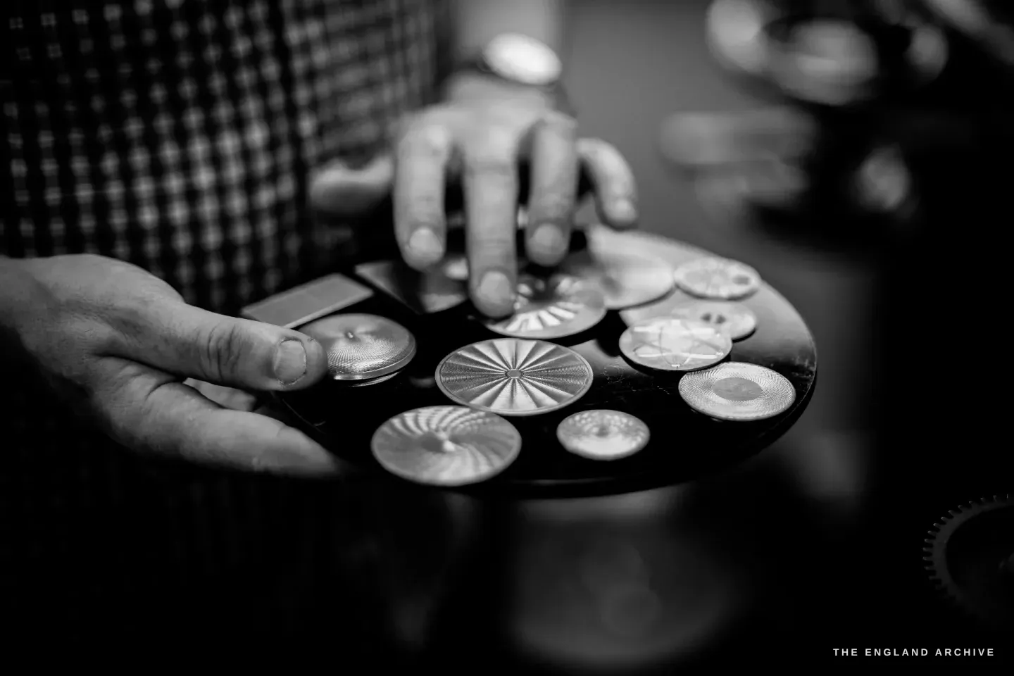 Hands holding a dark round tray of about ten engine-turned silver discs, each cut with a different pattern - sunburst, basketweave, spiral.