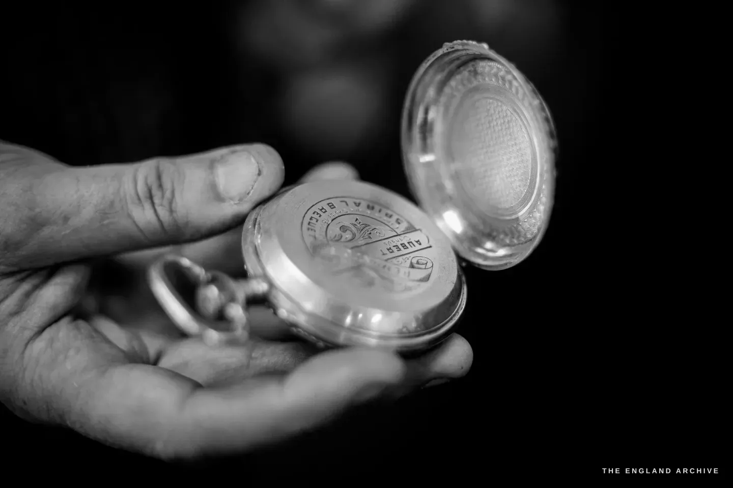 An open silver pocket-watch case held in a palm, the inner lid engine-turned, the engraving "SPIRAL BREGUET / AUBERT Genève" visible.
