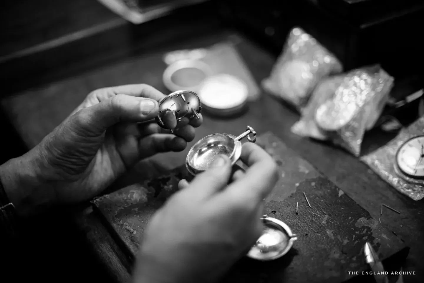 Two hands assembling a silver pocket-watch case at the bench, bagged parts and a finished watch to the right.