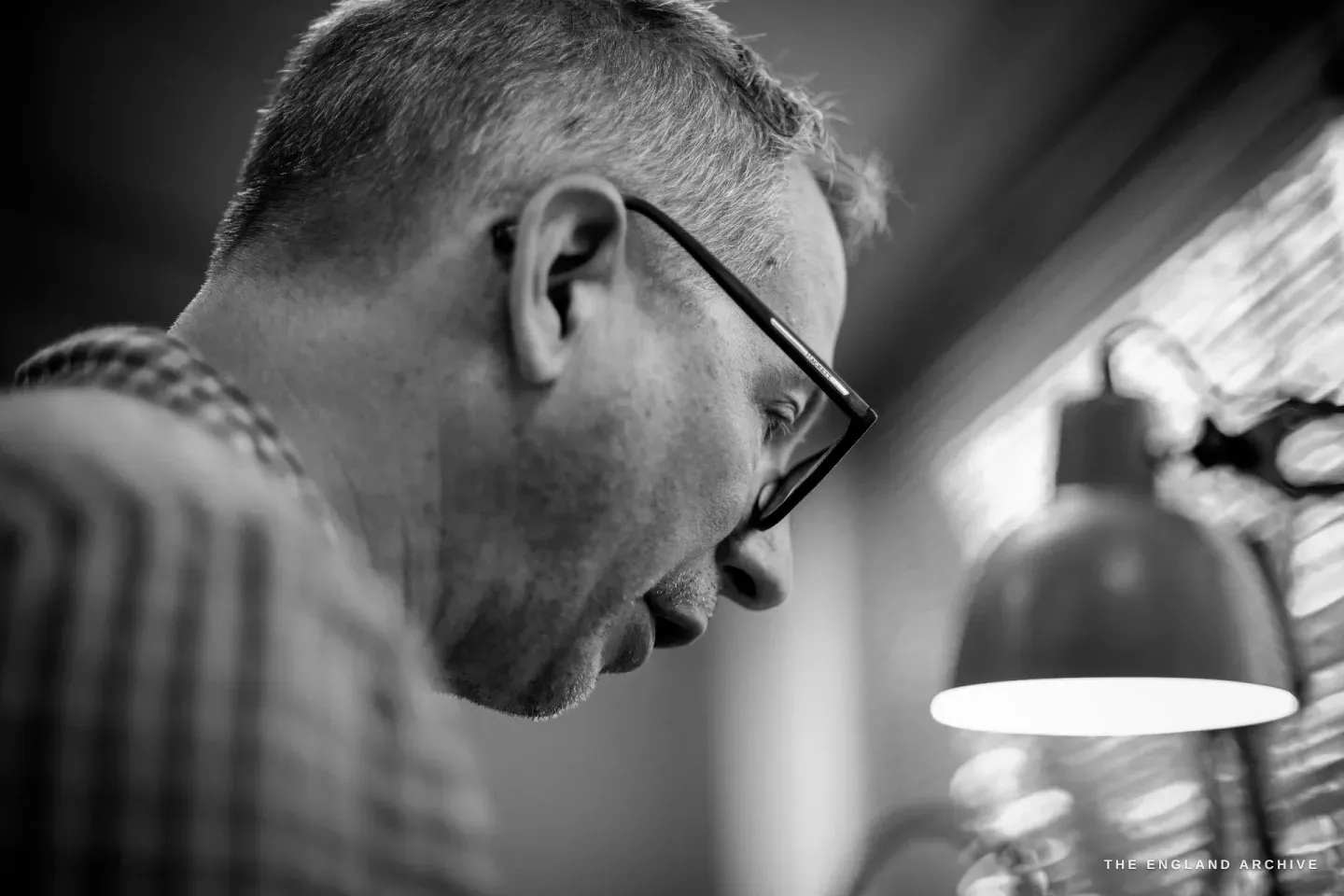 A close profile of Seth Kennedy in glasses looking down at his work, the bench lamp glowing beside him.
