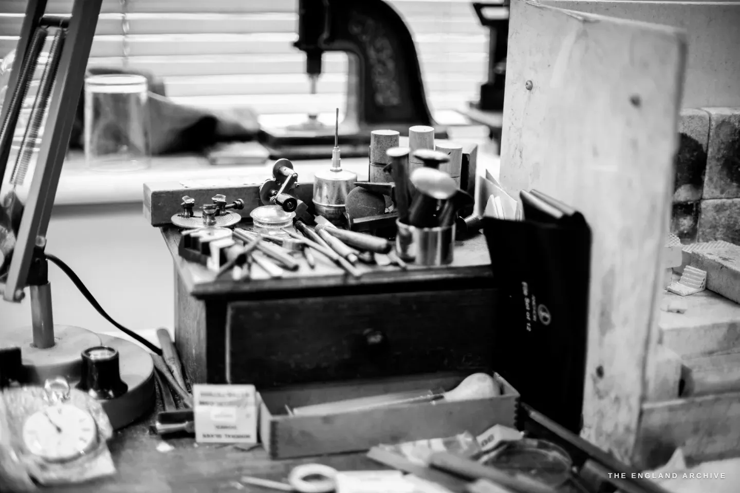 A bench still life - lathe tools and stakes in a wooden block on a drawer, a pocket watch at left, a brick wall behind.