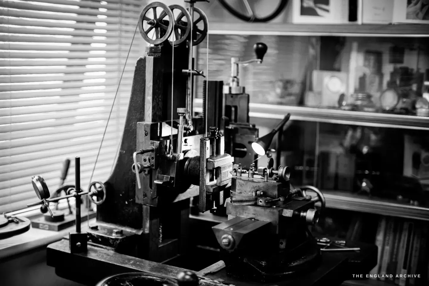 The upright straight-line engine-turning machine - twin pulley wheels at the top of a vertical column, a cross-slide below, set against window blinds.