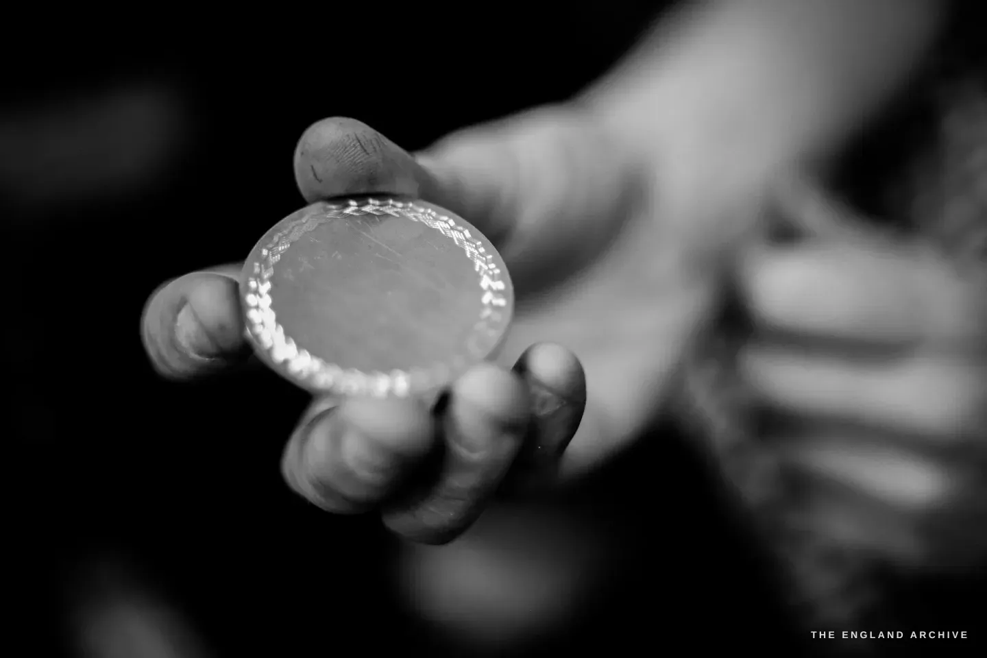 A hand holding a finished engine-turned silver disc up toward the camera, a second hand blurred behind.