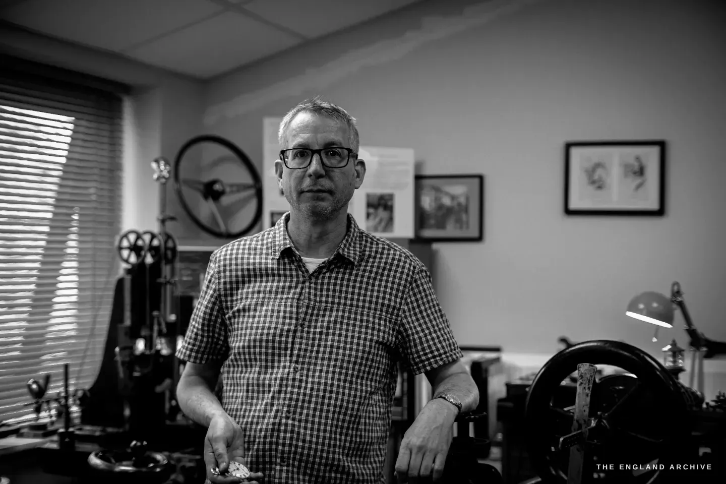 Seth Kennedy standing in the centre of his workshop holding a watch part, the upright straight-line engine to his left and the rose engine to his right.