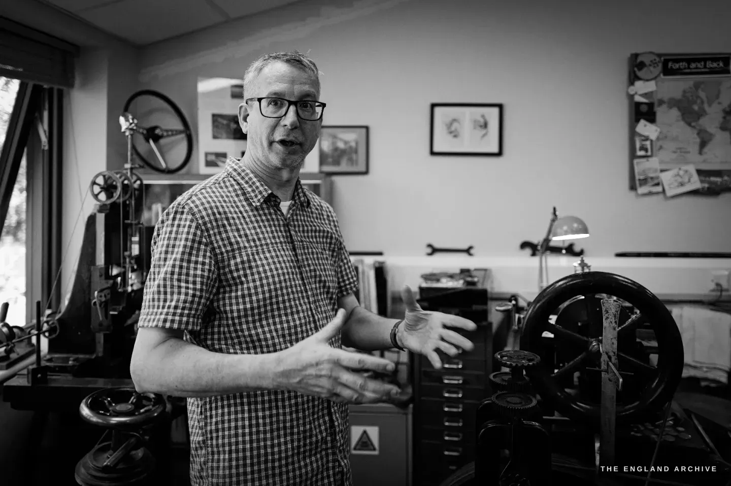 Seth Kennedy facing the camera with his hands open mid-explanation, the engine-turning machines to his left and a "Forth and Back" world map and framed prints on the wall behind.