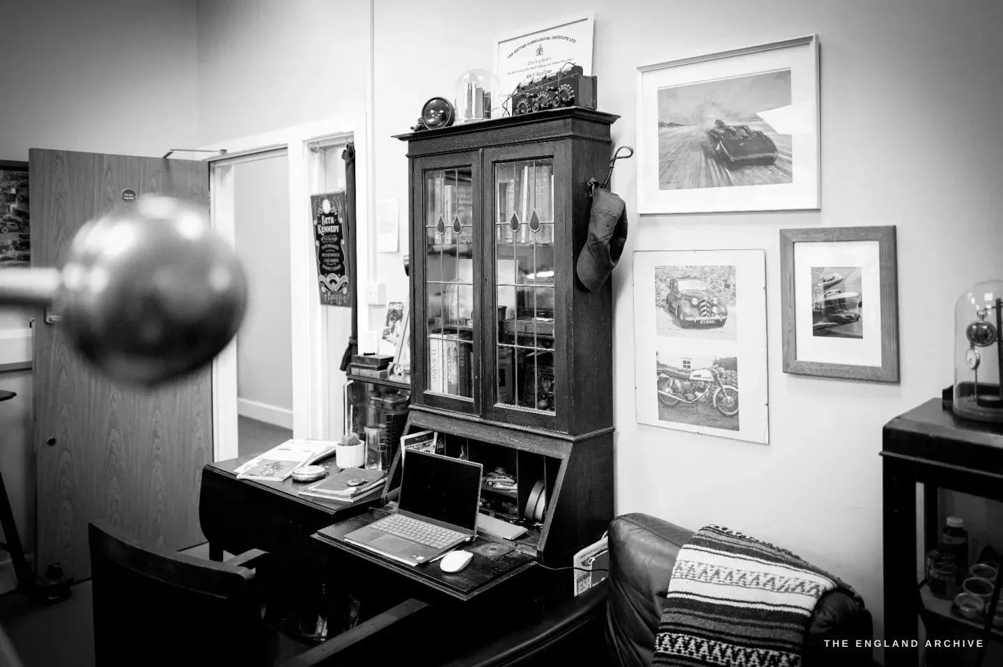 The office corner of the workshop - a glazed oak bookcase, framed vintage motoring prints, a laptop on a desk, a glass dome cloche and a leather chair.