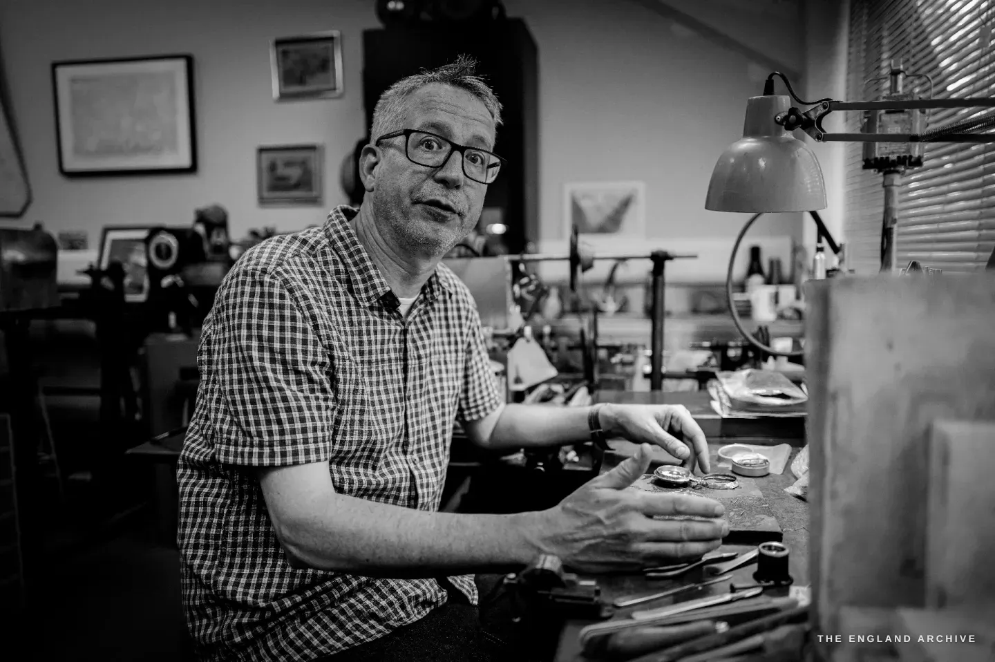 Seth Kennedy seated at his bench, looking to the camera with his hands resting over a watch part, the anglepoise lamp at his right and framed prints behind.