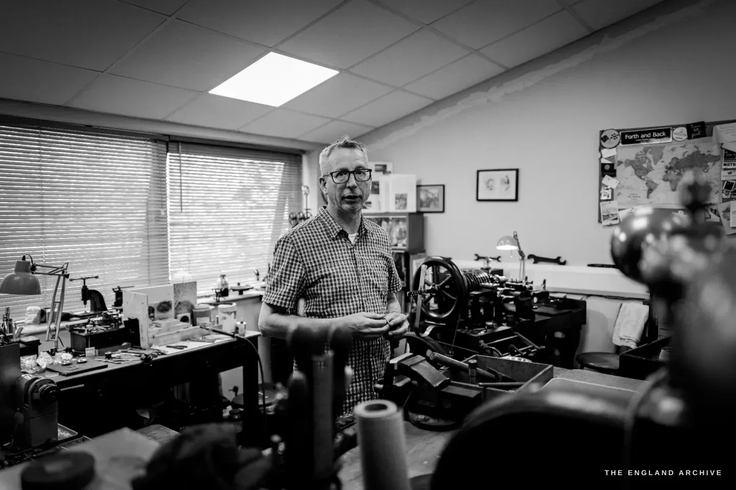 A wide view of the workshop with Seth Kennedy standing among the machines holding a part, blinds and a ceiling light above, the straight-line engine in the foreground.