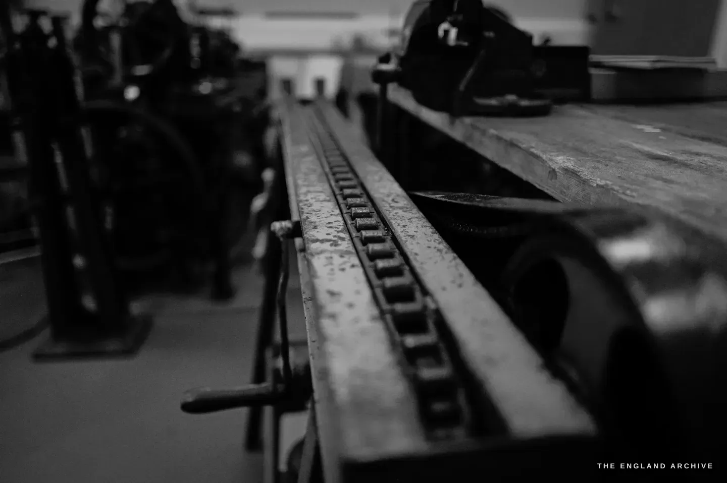 A low-angle view down the toothed bed of the straight-line engine, the crank handle at the far end, the workshop behind.