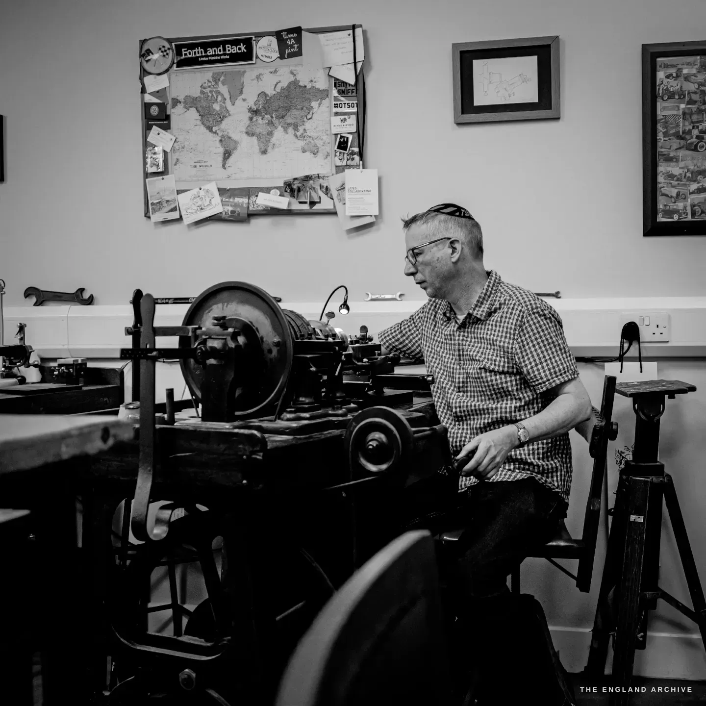 Seth Kennedy seated working the rose engine, a large dark flywheel at left, the "Forth and Back" map, framed prints and a wall spanner behind.