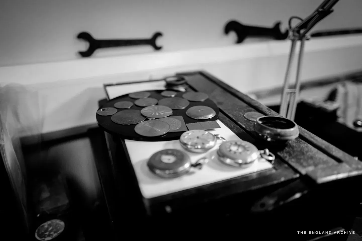 A bench still life - a dark tray of engine-turned discs and several silver pocket watches laid on white card, two wall-mounted spanners behind.