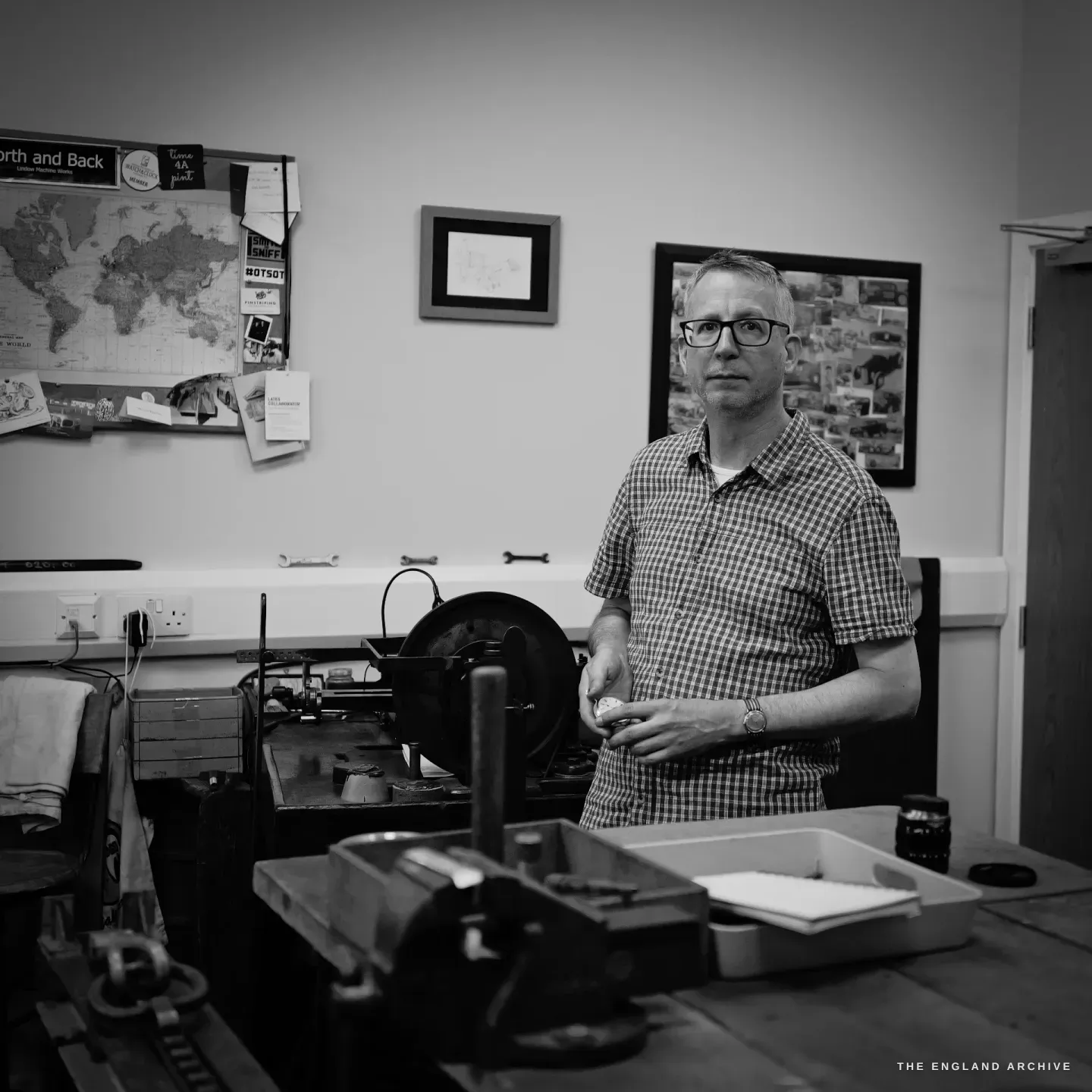 Seth Kennedy standing behind a bench with a vise, a part in hand, facing the camera, the "Forth and Back" world map and framed prints behind.