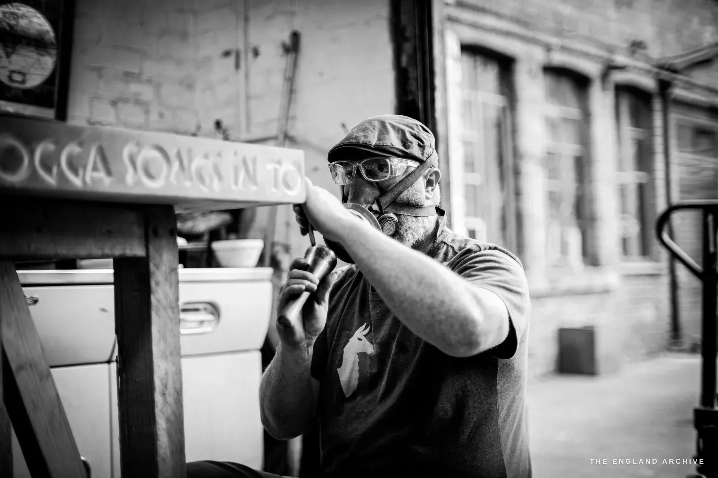 Steve Roche cutting the long stone sign out in the yard, a respirator over his mouth and safety goggles up on his cap, holding the lettered slab close to his face to check a cut, the words ‘songs in to…’ running along its edge.