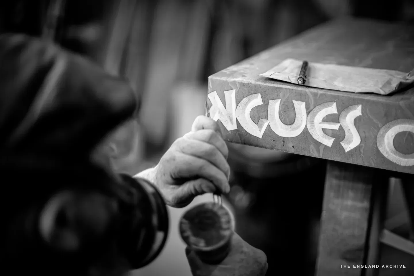A close study over Steve Roche’s shoulder of his hand driving a lettering chisel into the carved word ‘NGUES’ on a grey stone slab, a pencilled paper template and a pen resting on the stone above.