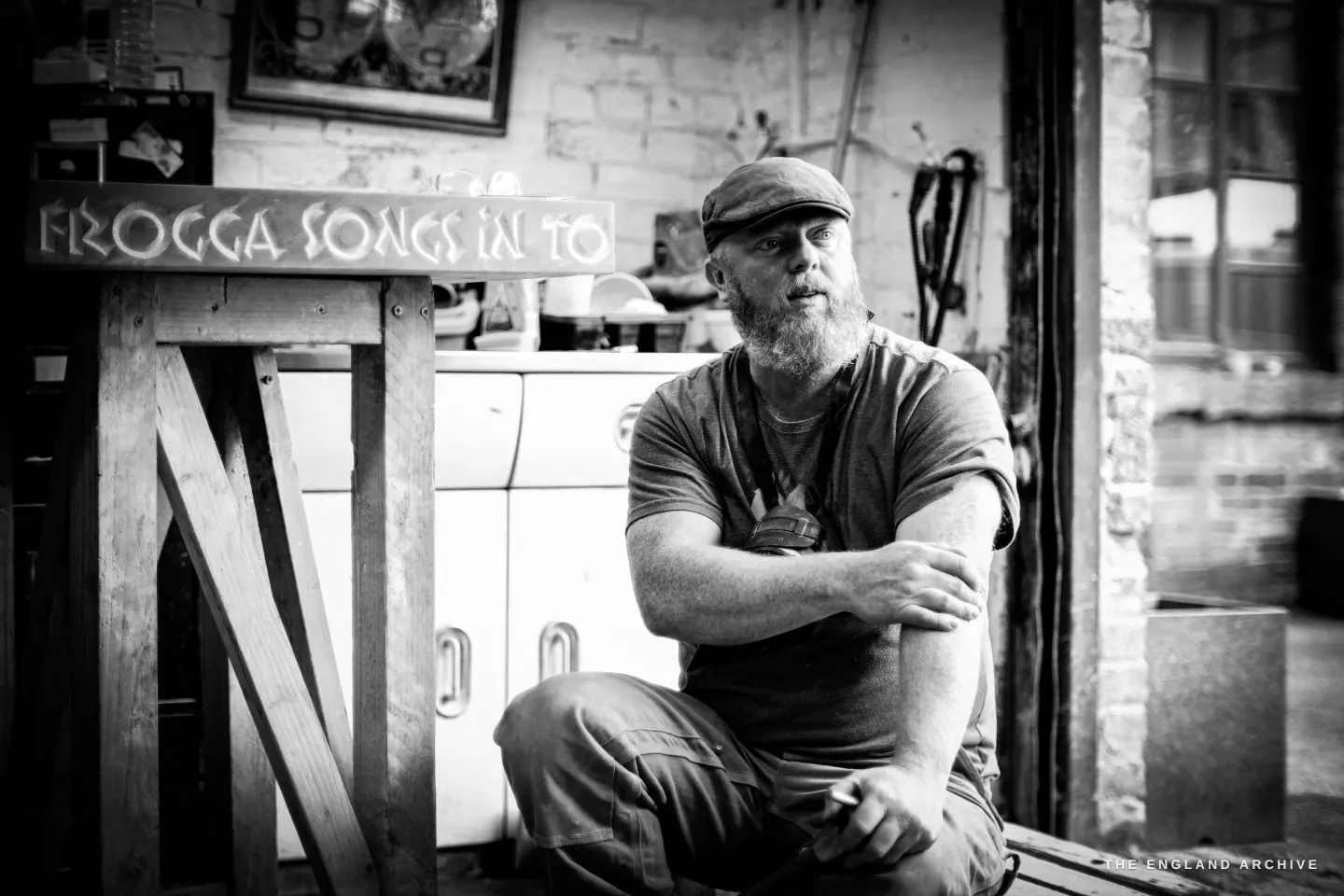 Steve Roche sitting on a low bench out in the works yard, arms folded over one knee, a flat cap and a full beard, looking off to one side; a long carved stone sign reading ‘songs in to…’ propped on a trestle behind him.