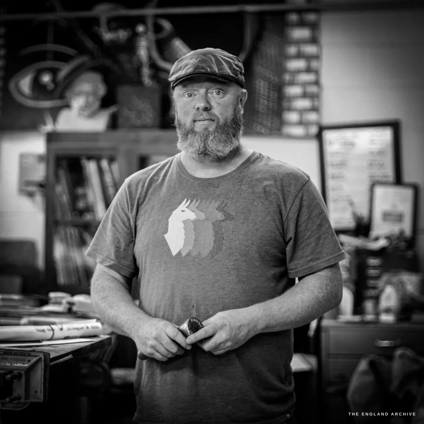 Steve Roche facing the camera in his workshop, flat cap and full beard, a grey t-shirt printed with a llama-and-stag motif, holding a small tin of tooling wax, the studio behind him.
