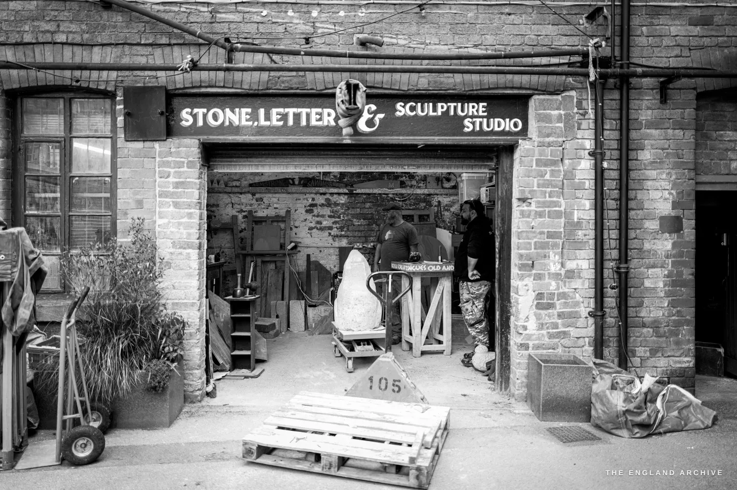 The street frontage of the Stone, Letter and Sculpture Studio at Stag Works in Sheffield - a brick archway beneath a hand-painted sign, a half-carved white stone on a trolley just inside the open doorway, a pallet and a sack truck on the ground outside.