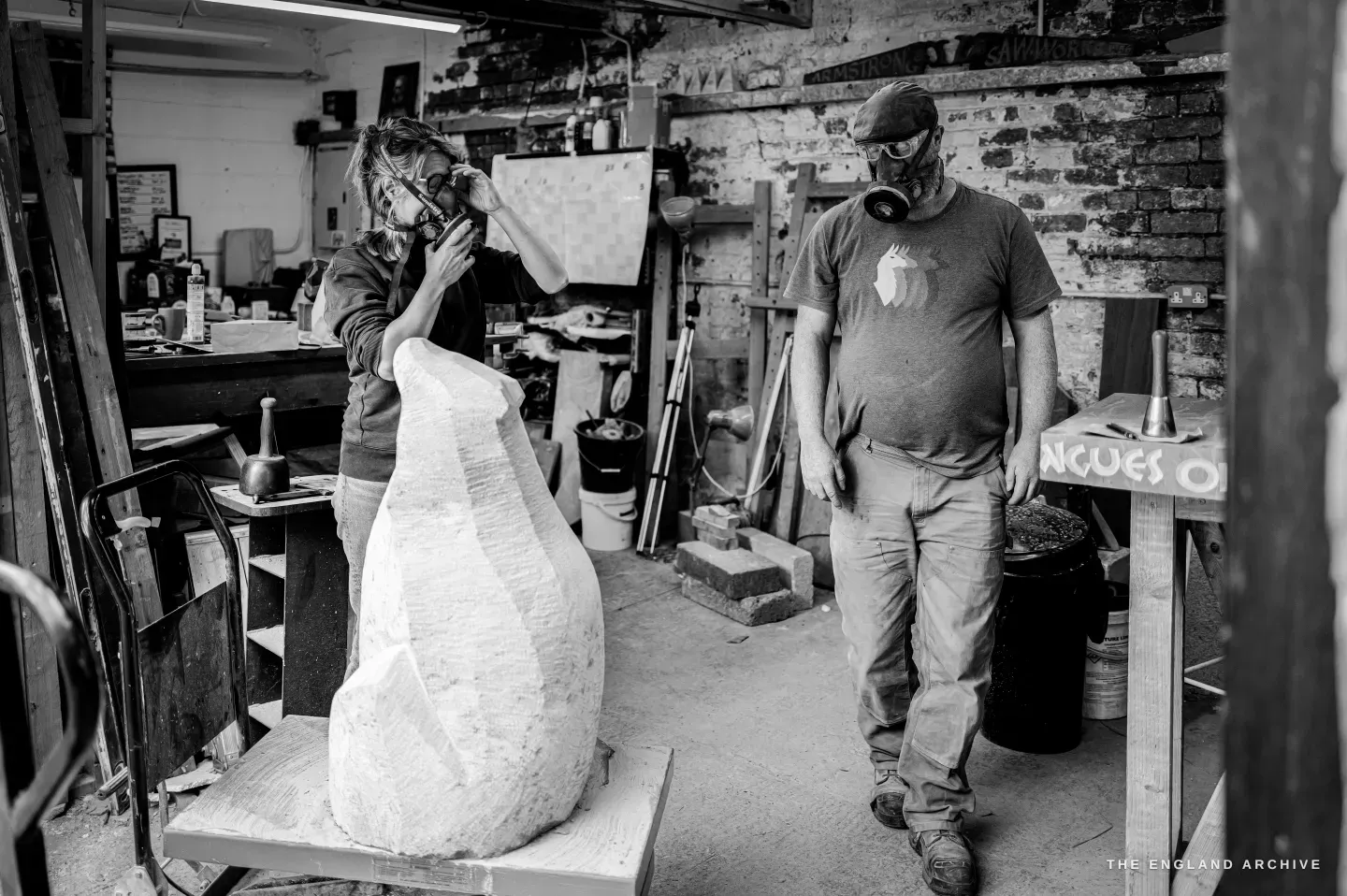 Lily Marsh in a respirator working at her roughed-out stone while Steve Roche stands a few feet away, hands at his sides, watching the work, the ‘songs in tongues’ sign on the bench behind them.