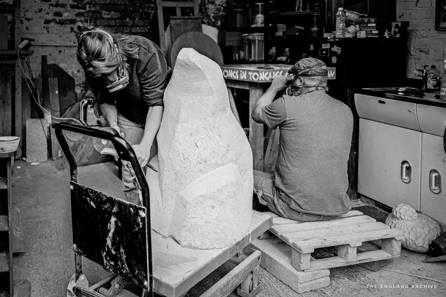 Lily Marsh bent over her sculpture with a chisel while Steve Roche works at the sink-side bench with his back to the camera, a small carved stone sheep sitting on a wooden pallet on the floor between them.