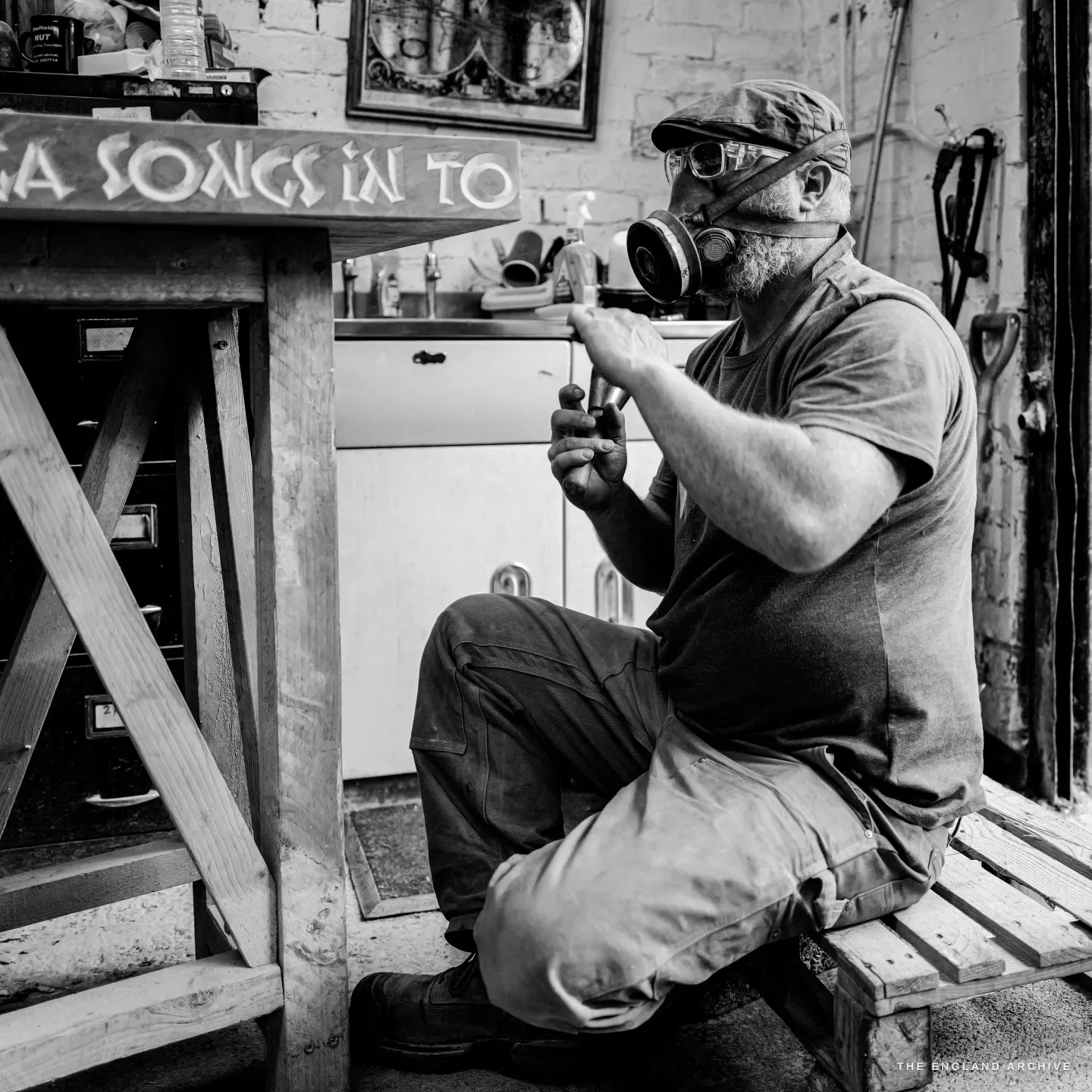 Steve Roche seated in the works yard in a respirator and safety goggles, a lettering chisel and a round dummy mallet held ready in his hands, the carved ‘songs in to…’ sign on a trestle beside him.