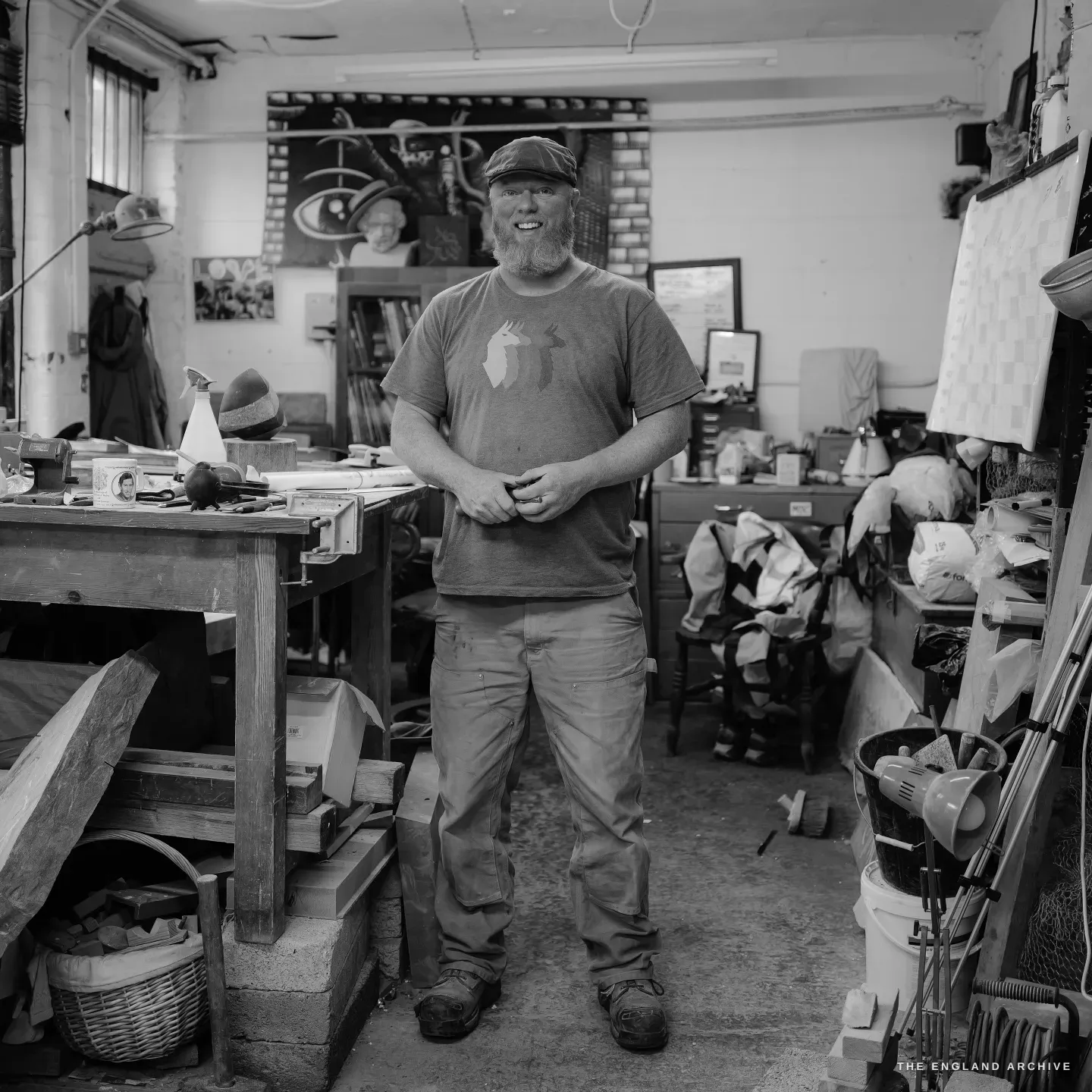 Steve Roche standing in the middle of the shared studio, hands clasped in front of him, a slight smile, the cluttered workshop and a large eye-and-antlers artwork on the wall behind him.