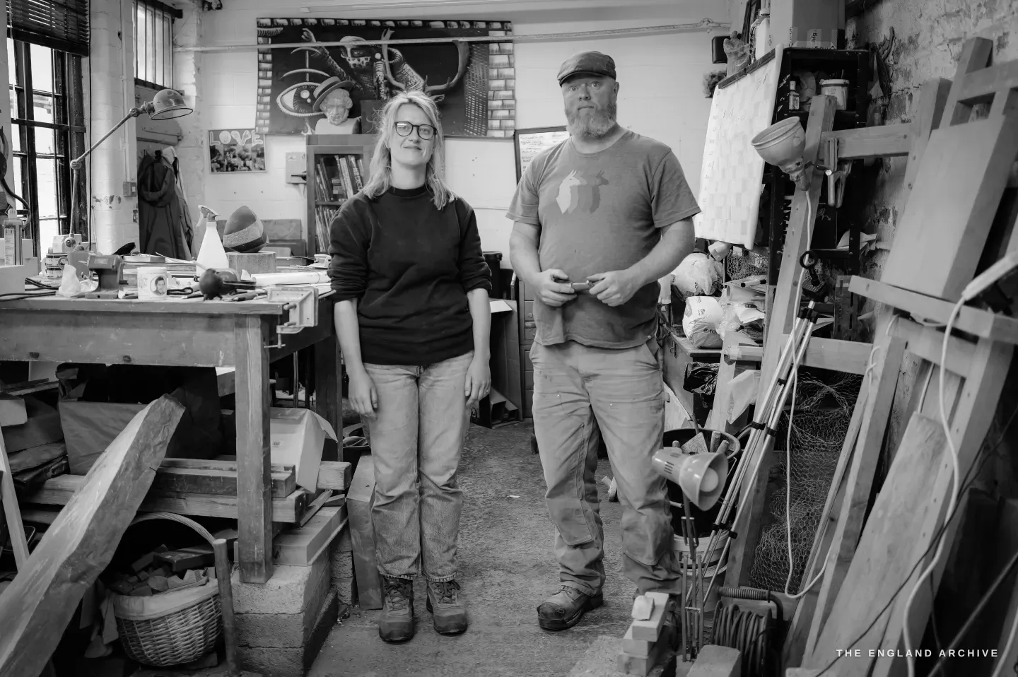 Steve Roche and Lily Marsh standing together in the middle of the workshop facing the camera - Steve at right with a full beard and cap, Lily at left in a dark jumper, the bench and racked stone slabs around them.