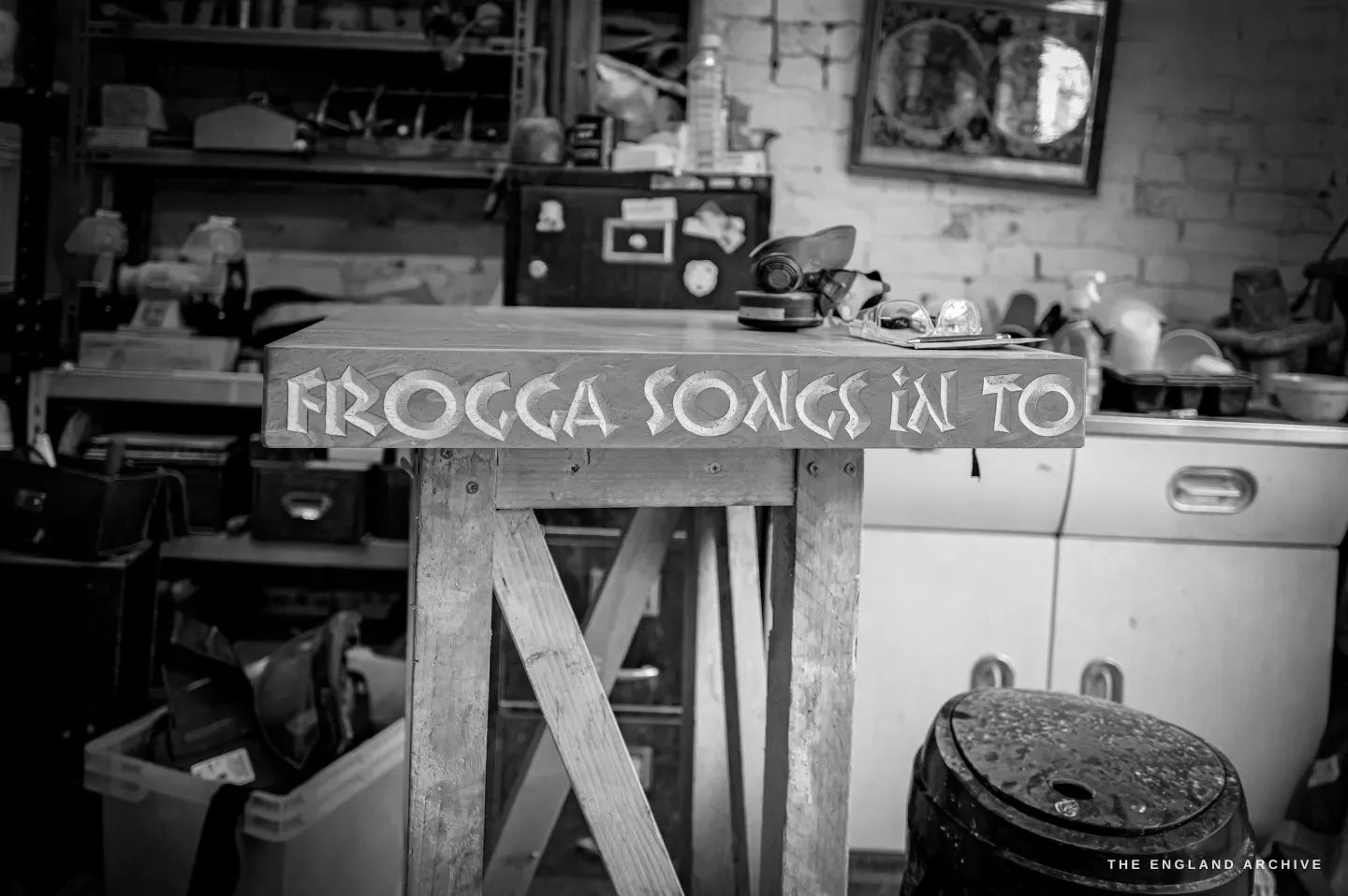 The finished carved sign resting on a trestle in the workshop, the lettering reading ‘Frogga songs in to…’ cut and painted into the long grey stone, the cluttered studio softened behind it.
