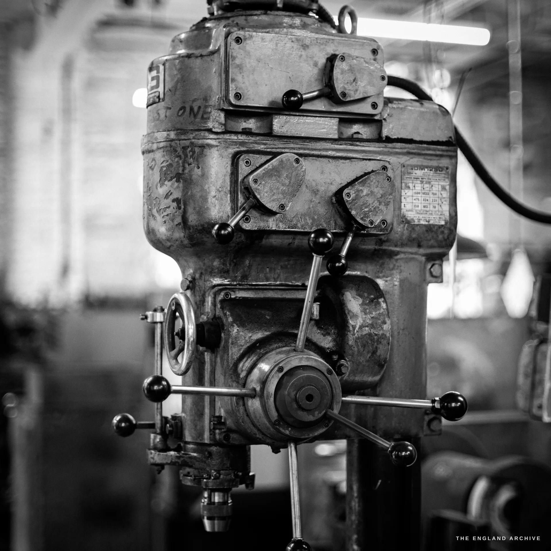 A close view of an old grimy machine tool on the factory floor - a vertical drilling head with hand levers and gears and a worn maker’s plate.