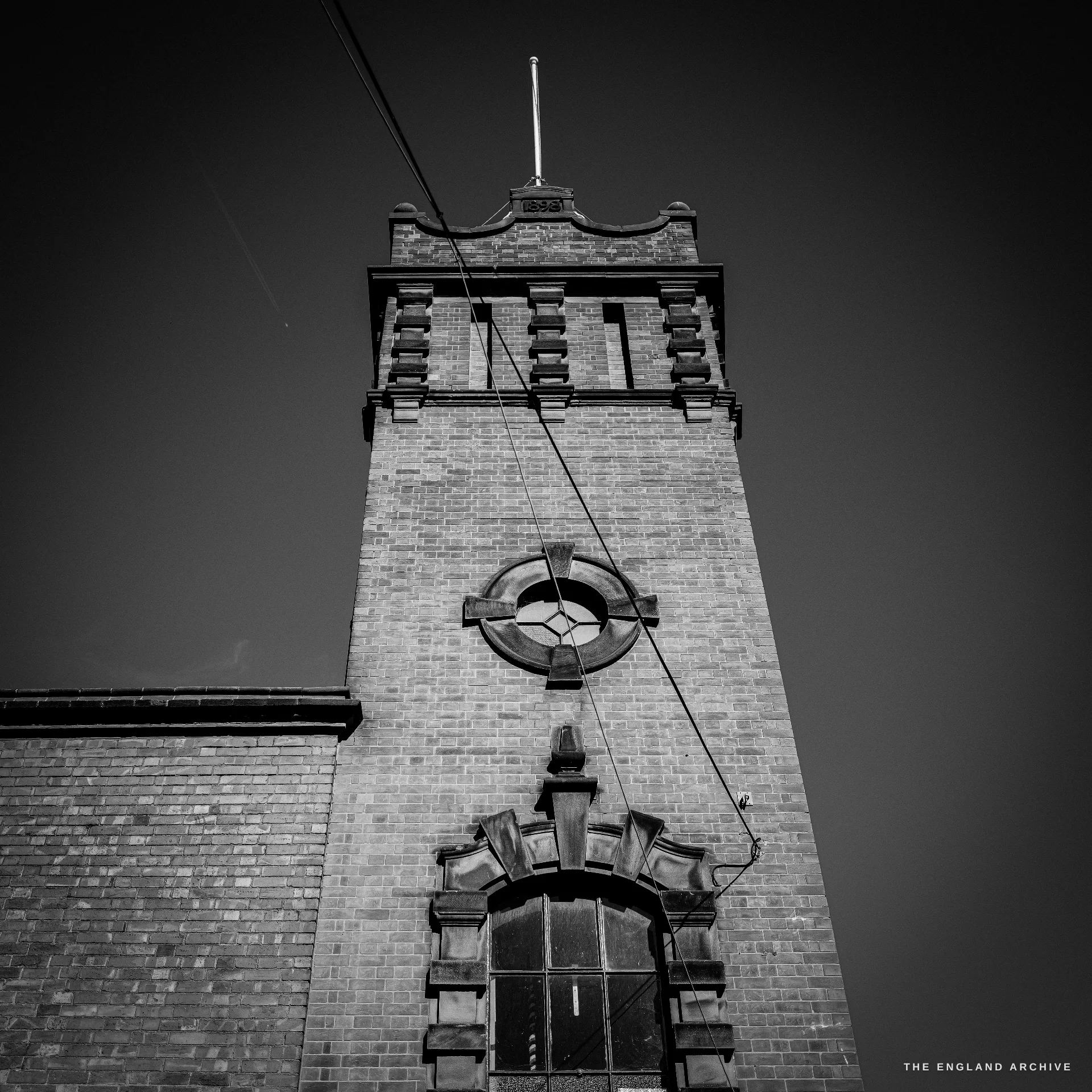 Looking up at the foundry’s brick clock-and-bell tower with a circular vent, decorative brick cornice and a flagpole, overhead wires crossing the frame.