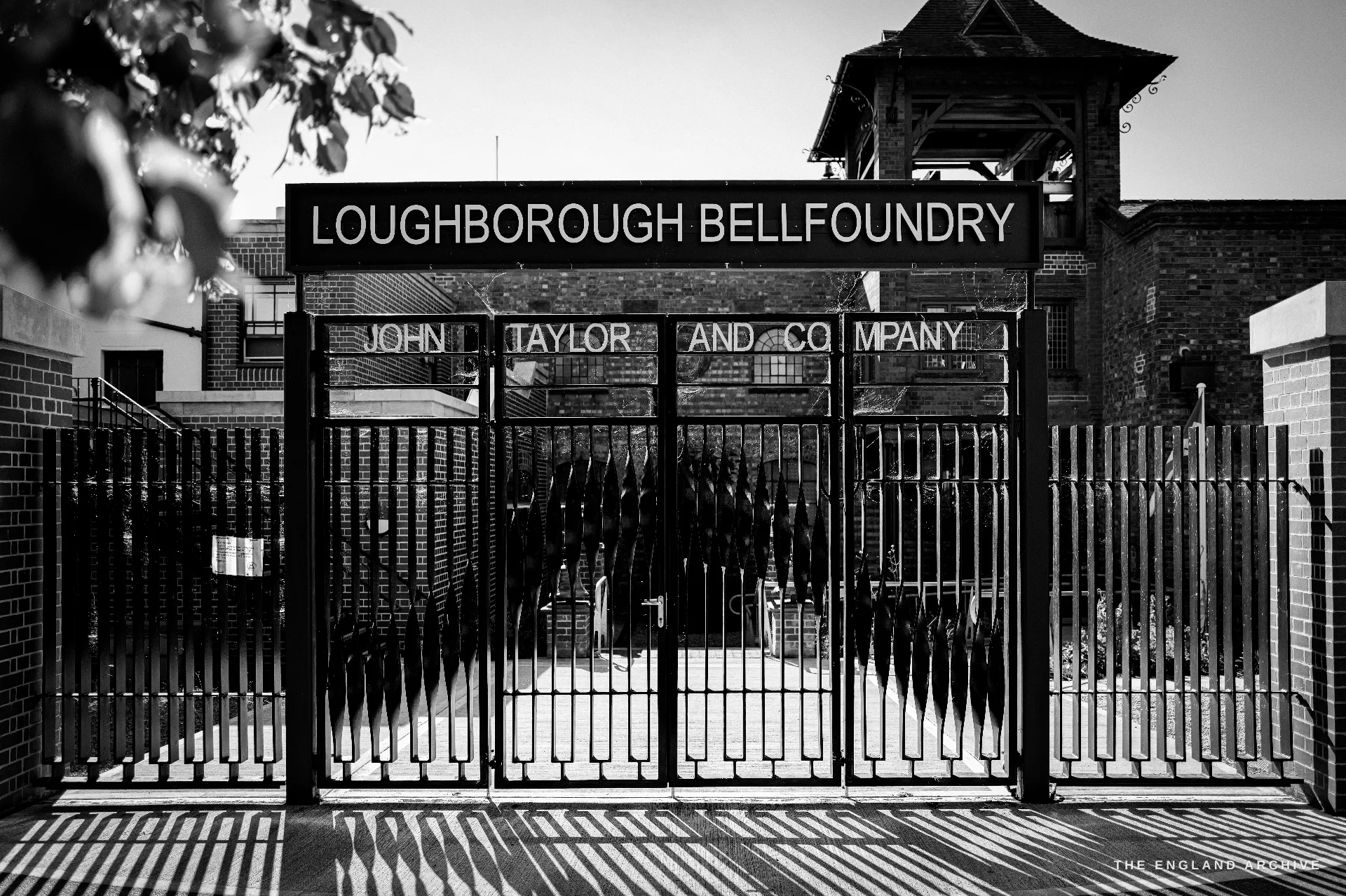 The main entrance gates of the works, black railings beneath a large lettered signboard reading LOUGHBOROUGH BELLFOUNDRY, JOHN TAYLOR AND COMPANY, the bell tower behind.