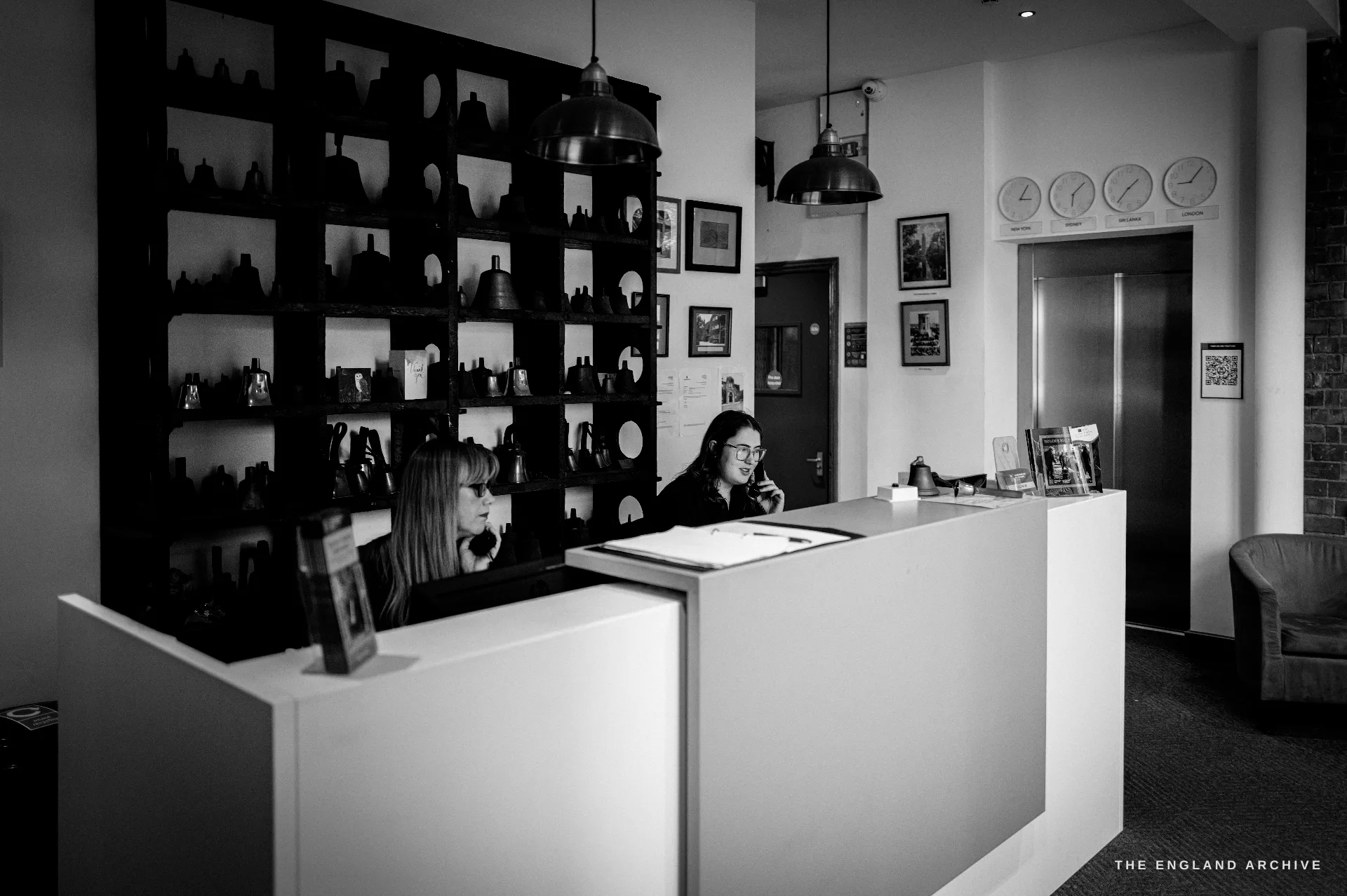 The museum reception foyer: two staff at a white desk, a tall dark shelving unit of bells behind, pendant lamps and a wall of clocks and framed photographs.