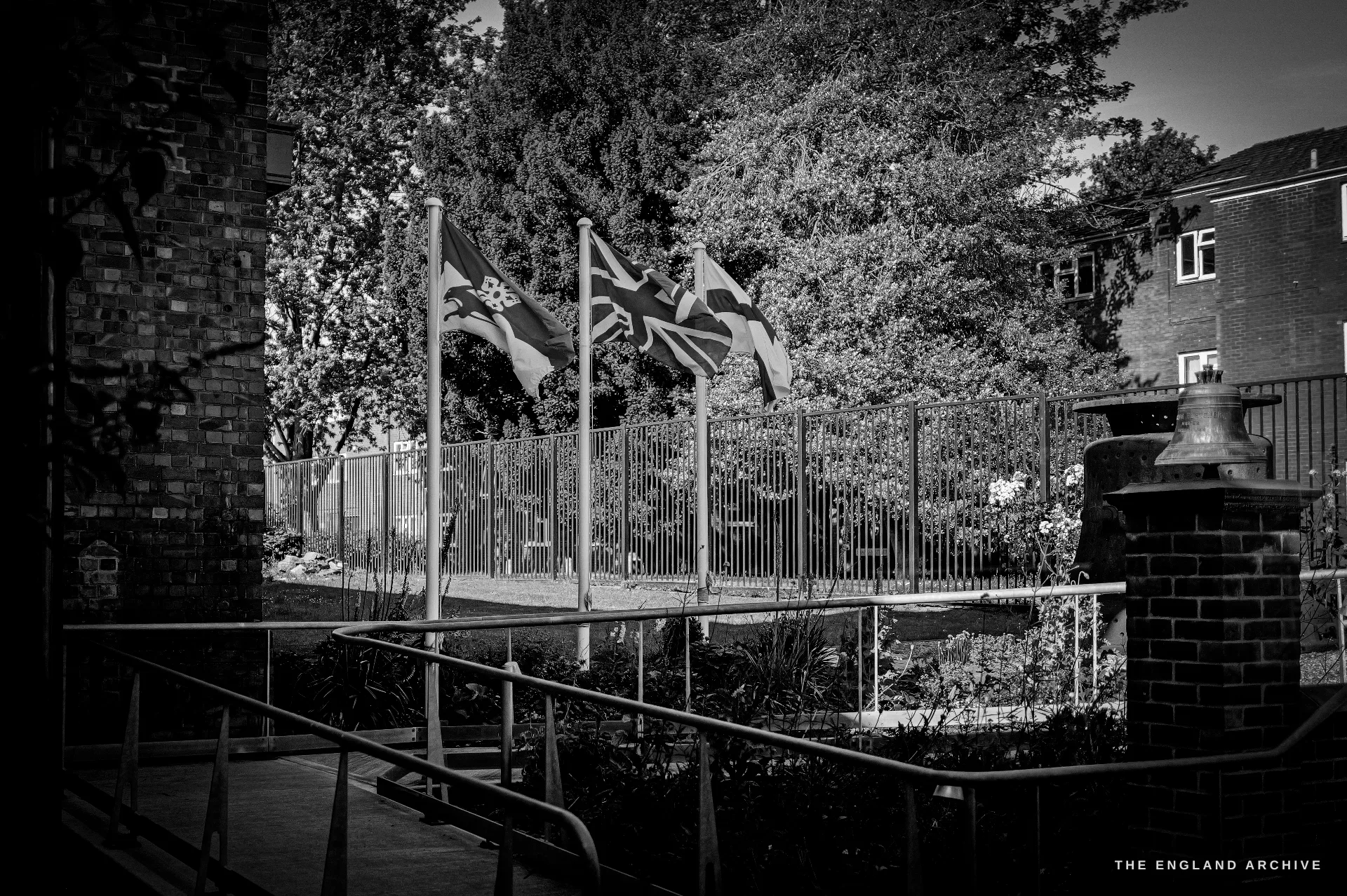 The museum garden and courtyard with three flags flying over a railed planted area, a brick plinth and bell at right, trees behind.