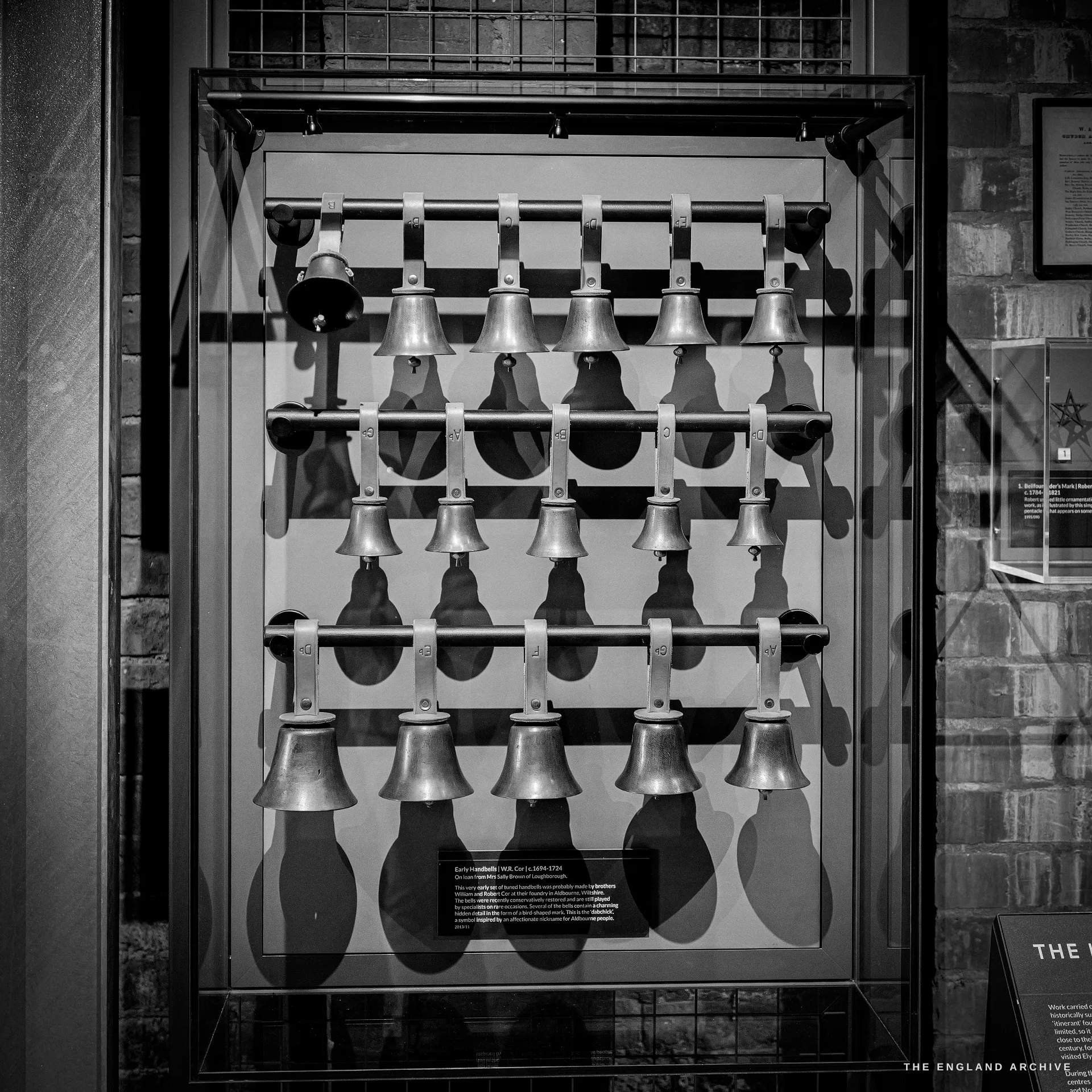 A glazed display case holding a graduated, tiered rack of handbells mounted in rows, with an interpretation card, the brick room reflected in the glass.