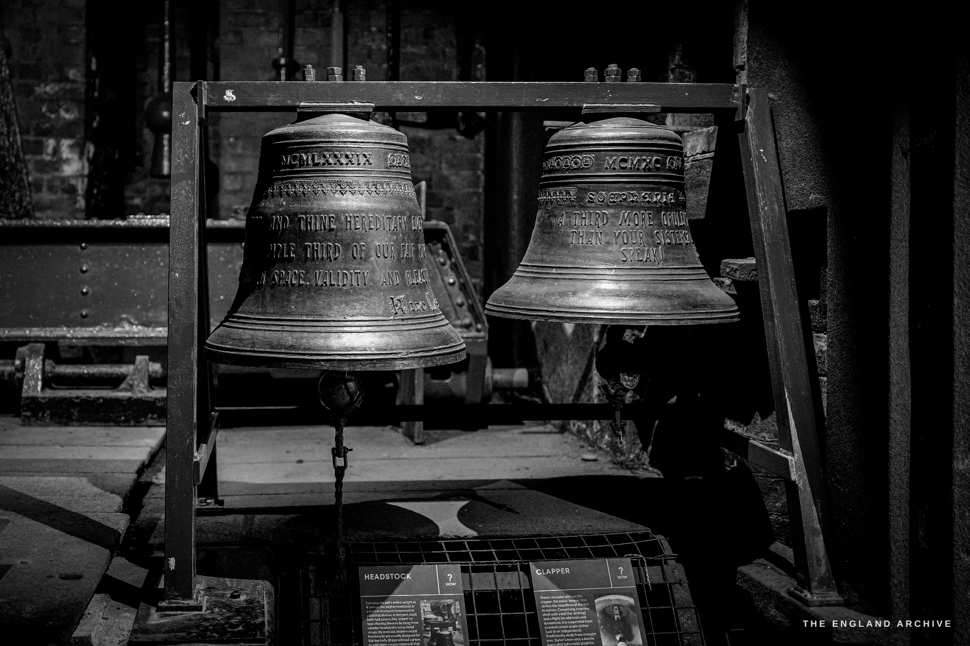 Two large bronze bells hung side by side on a frame as a museum exhibit, cast inscriptions and Roman-numeral dates around their shoulders.