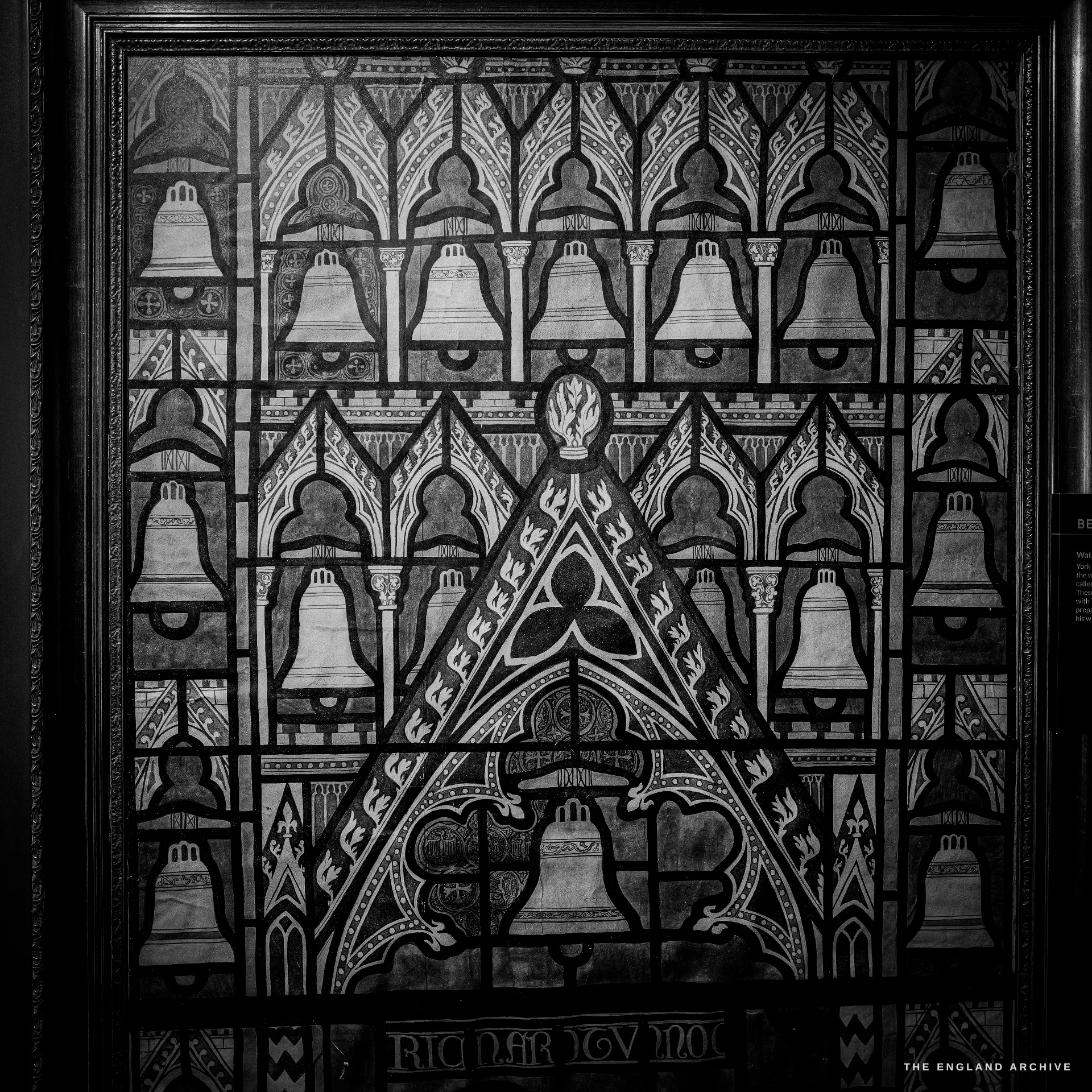 A framed stained-glass panel depicting rows of bells in a Gothic pattern, displayed on a dark museum wall.