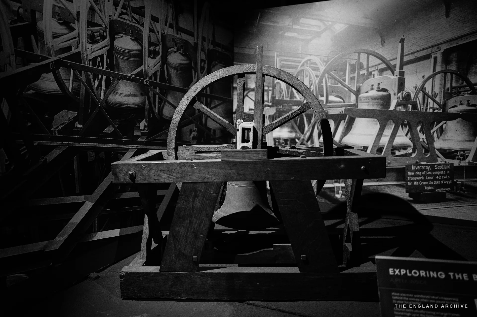 A museum exhibit of a complete bell-hanging assembly - a bell mounted in a wooden frame with its large wheel and headstock - against a painted mural of a belfry.