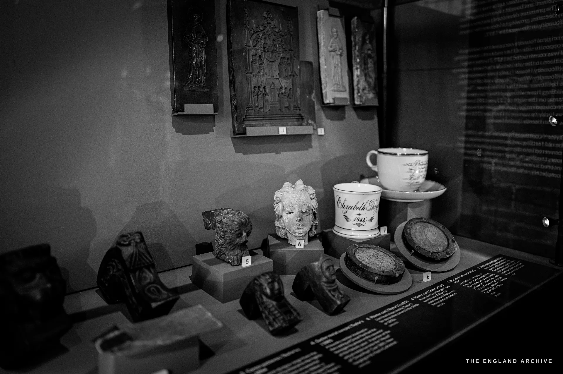 A glazed display case of small artefacts - cast relief fragments, a moulded classical head, inscribed commemorative mugs and plaques - with caption cards.
