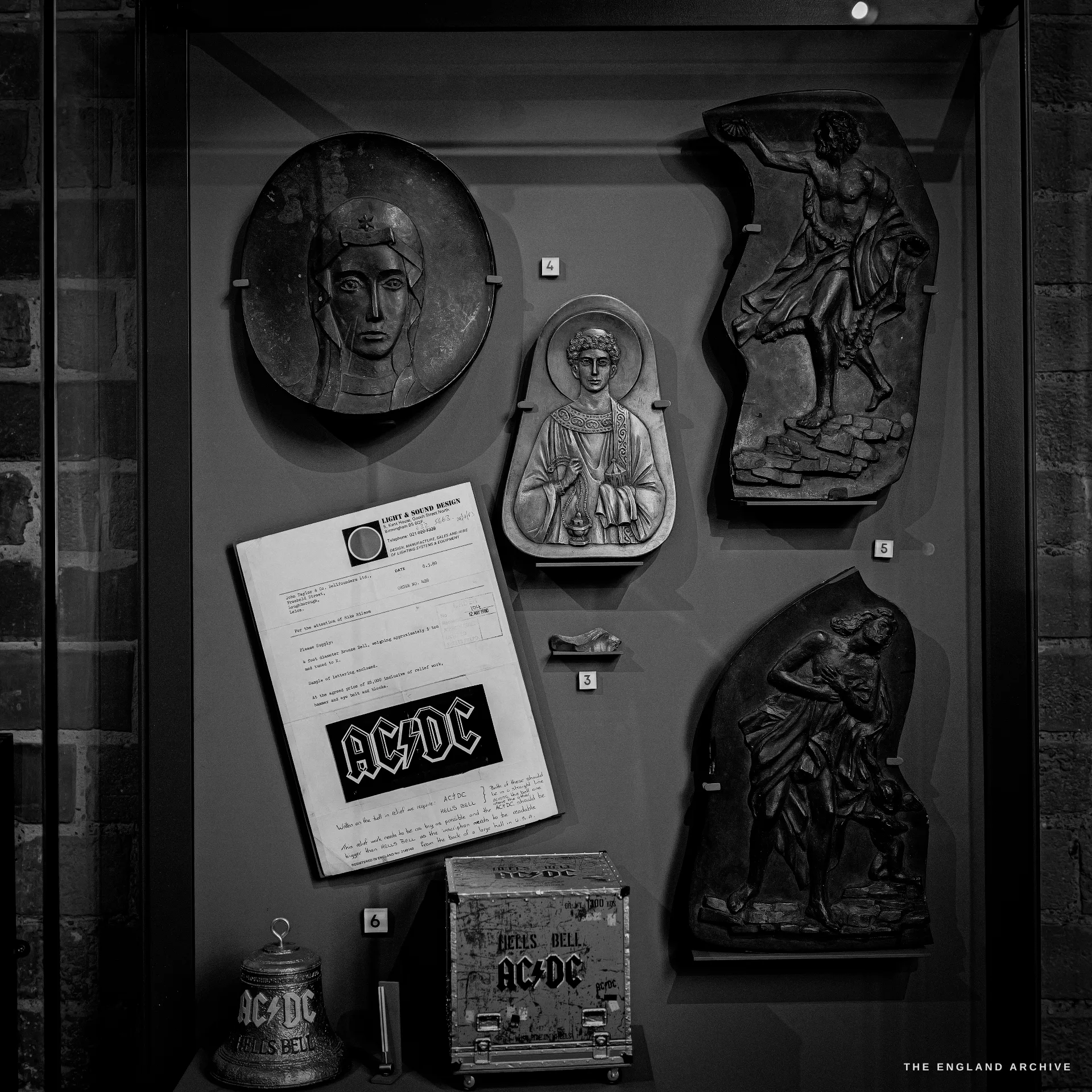 A display case of decorative bell-casting reliefs and patterns alongside AC/DC Hells Bells memorabilia and a framed document.