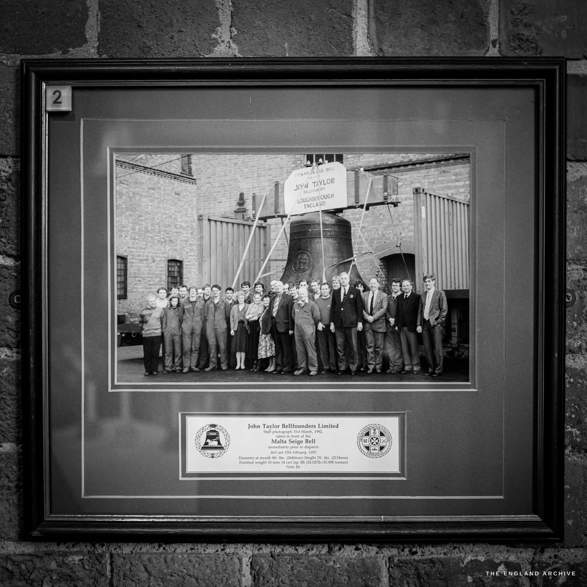 A framed historic black-and-white photograph of the foundry workforce posed in front of a very large bell in the yard, with a descriptive plaque below.