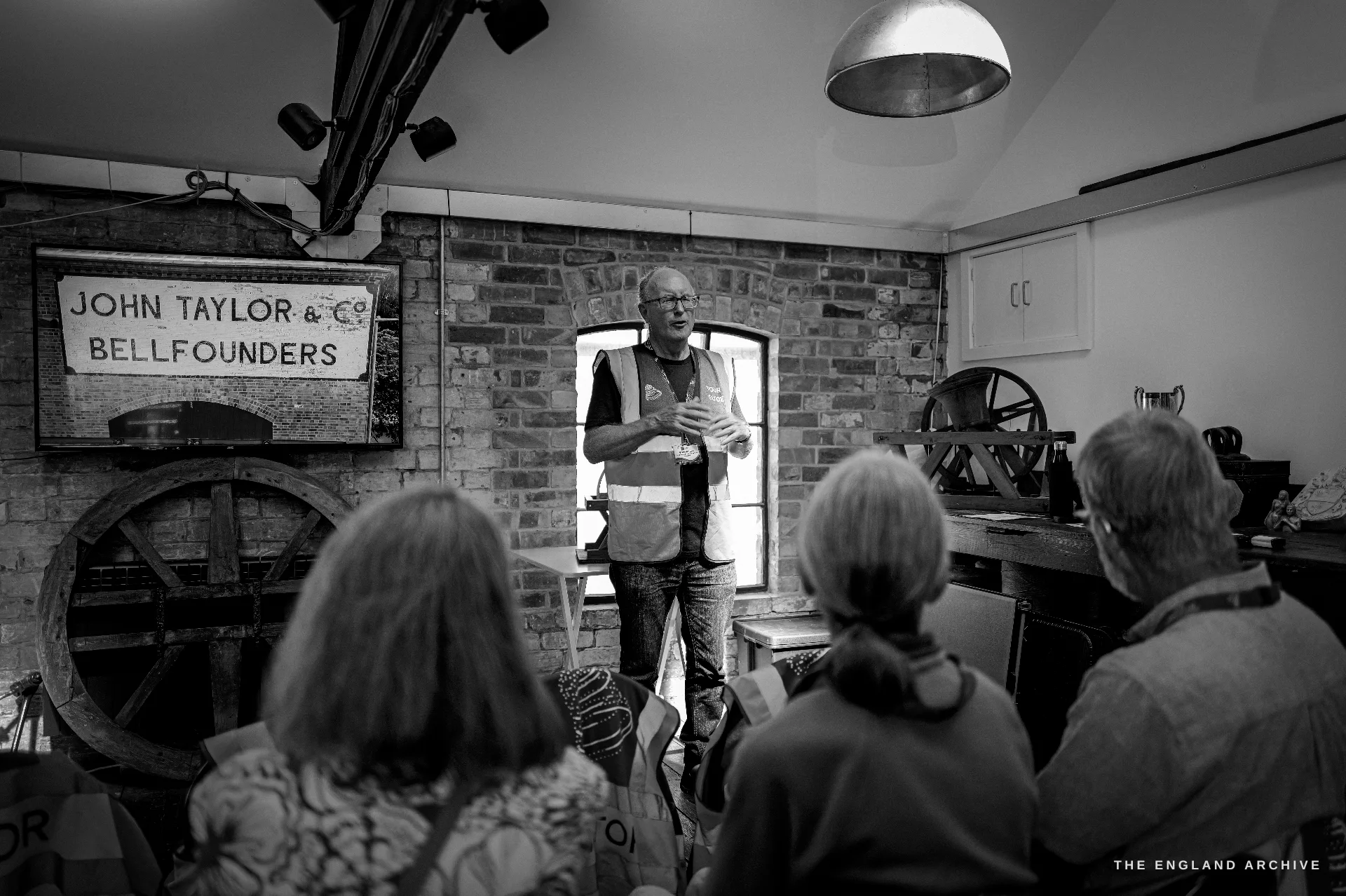 A tour guide in a hi-vis vest talking to a seated group of visitors in a brick room, a framed JOHN TAYLOR & Co sign and a wooden bell wheel on the wall behind.