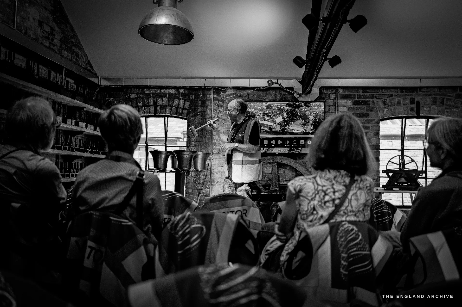 The same tour guide demonstrating a handbell to the seated audience, the backs of visitors’ heads in the foreground, exhibits shelved behind.