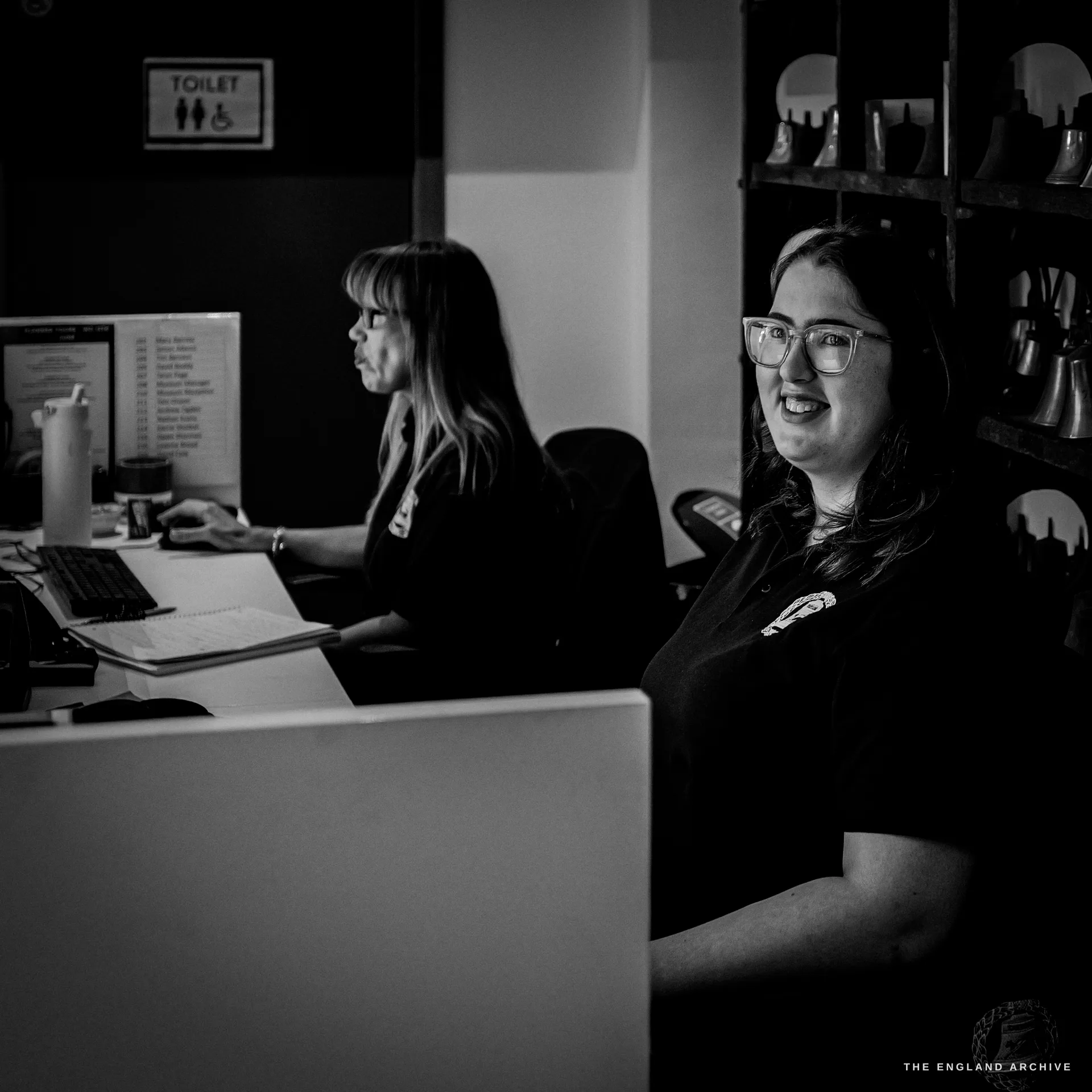 Two members of the reception team at the front desk, one at a computer and one turned and smiling, shelves of small bells behind them.