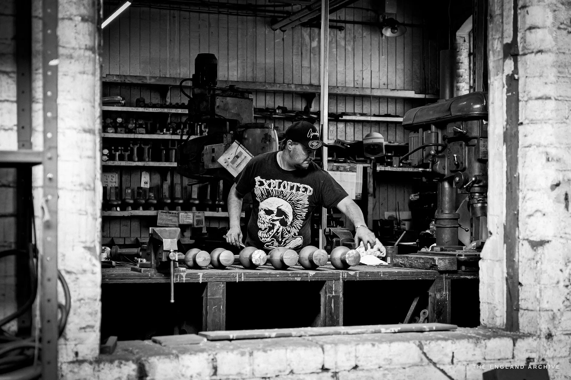 A worker in a black band t-shirt and cap working at a bench lined with turned clapper balls, seen through a doorway with tool racks behind.