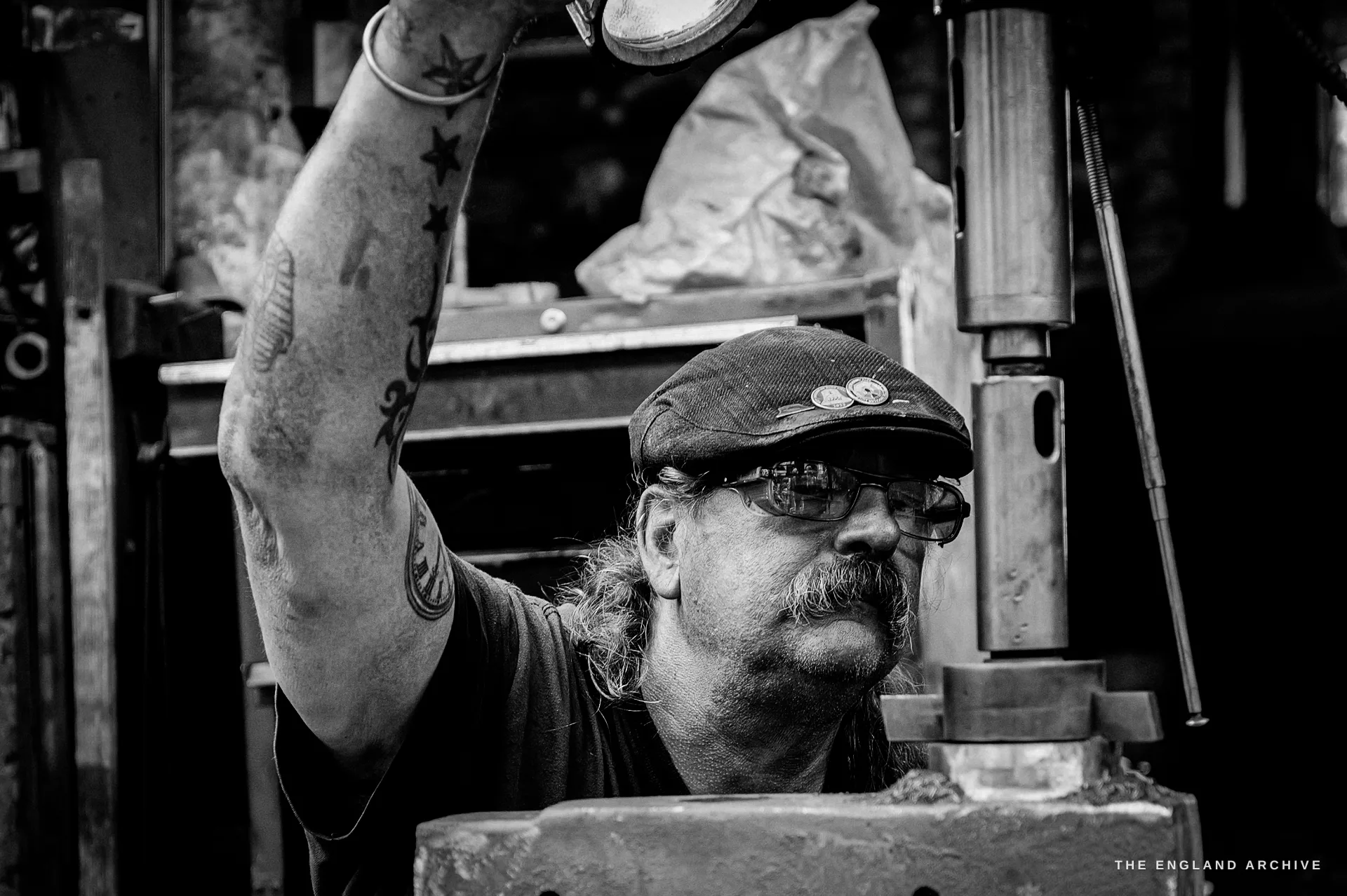 A close view of an older foundry worker in glasses, a moustache and a badged flat cap, his tattooed arm raised to the machinery.