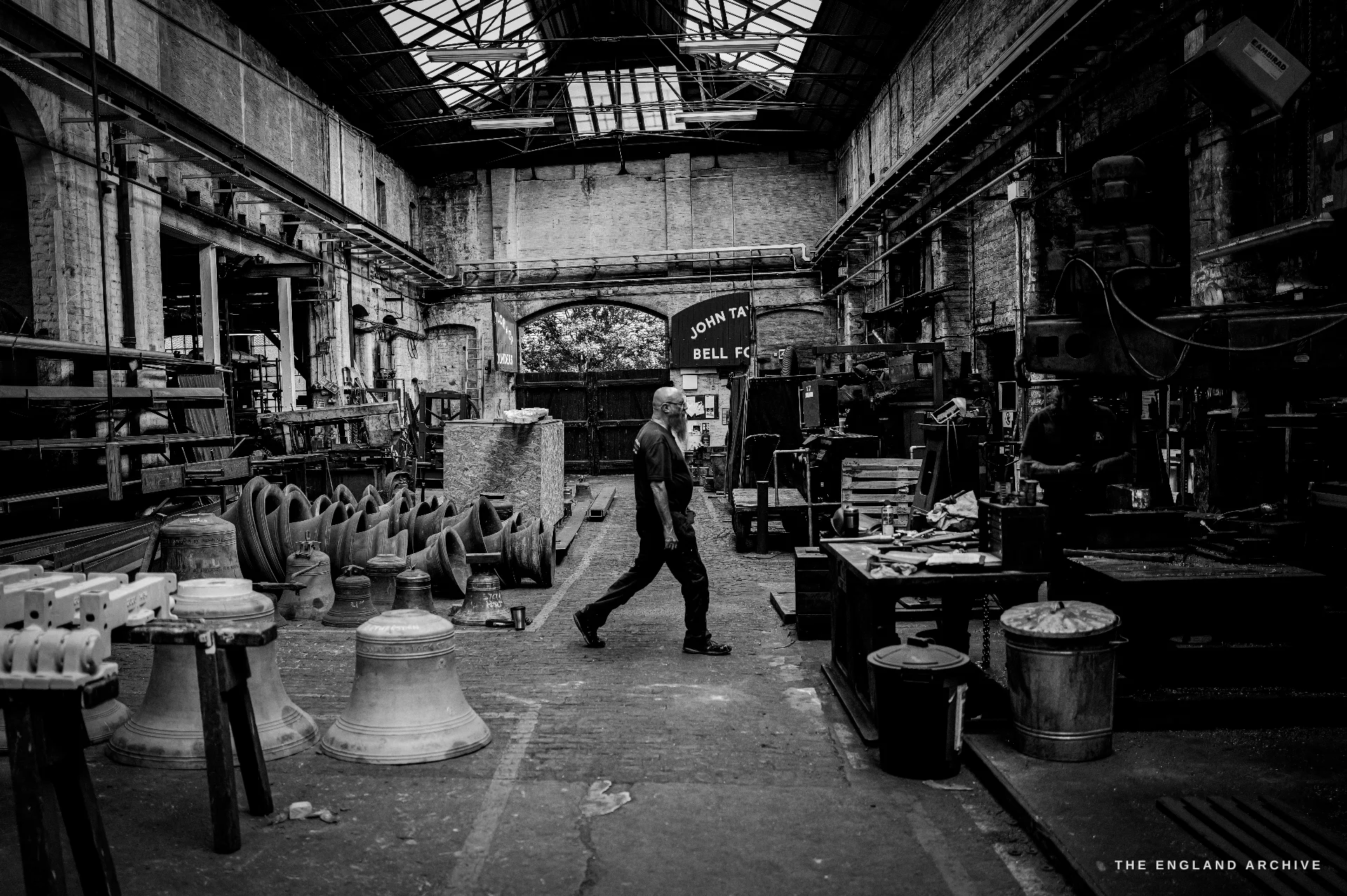 A wide view of the main casting hall - a worker crossing the floor, raw bells and moulds at left, benches at right, an arched skylit roof and a painted JOHN TAYLOR BELL FOUNDRY sign on the rear wall.