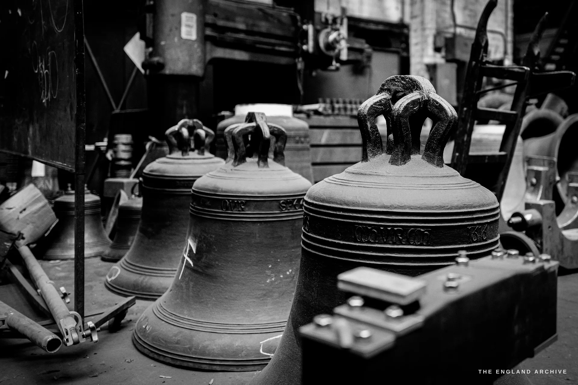 Three newly cast bronze bells with crown loops standing on the foundry floor, cast inscriptions around their shoulders, machinery and a barrow behind.