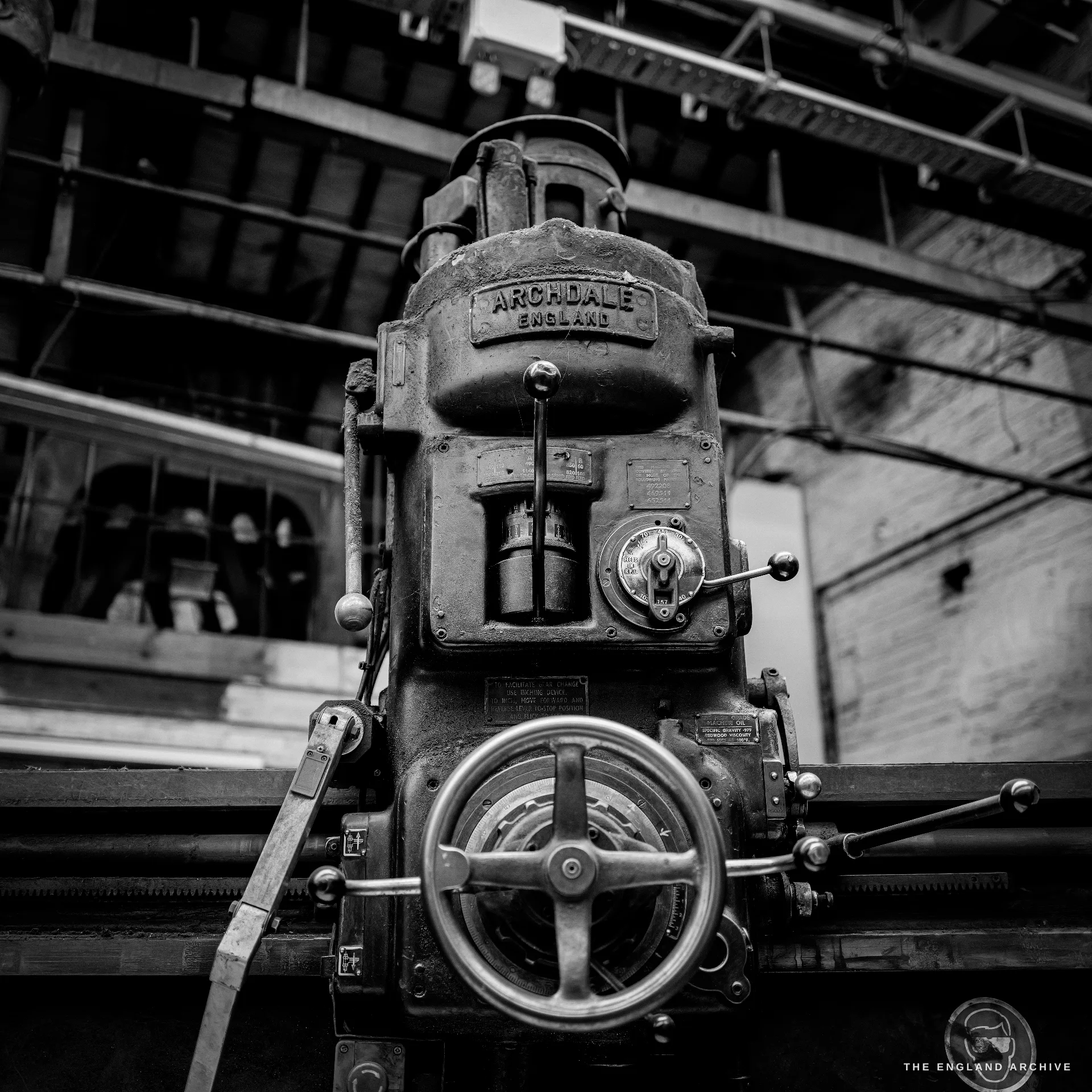 A heavy vintage radial drilling machine with a hand-wheel and a cast ARCHDALE ENGLAND maker’s plate, figures dimly visible through the workshop windows behind.